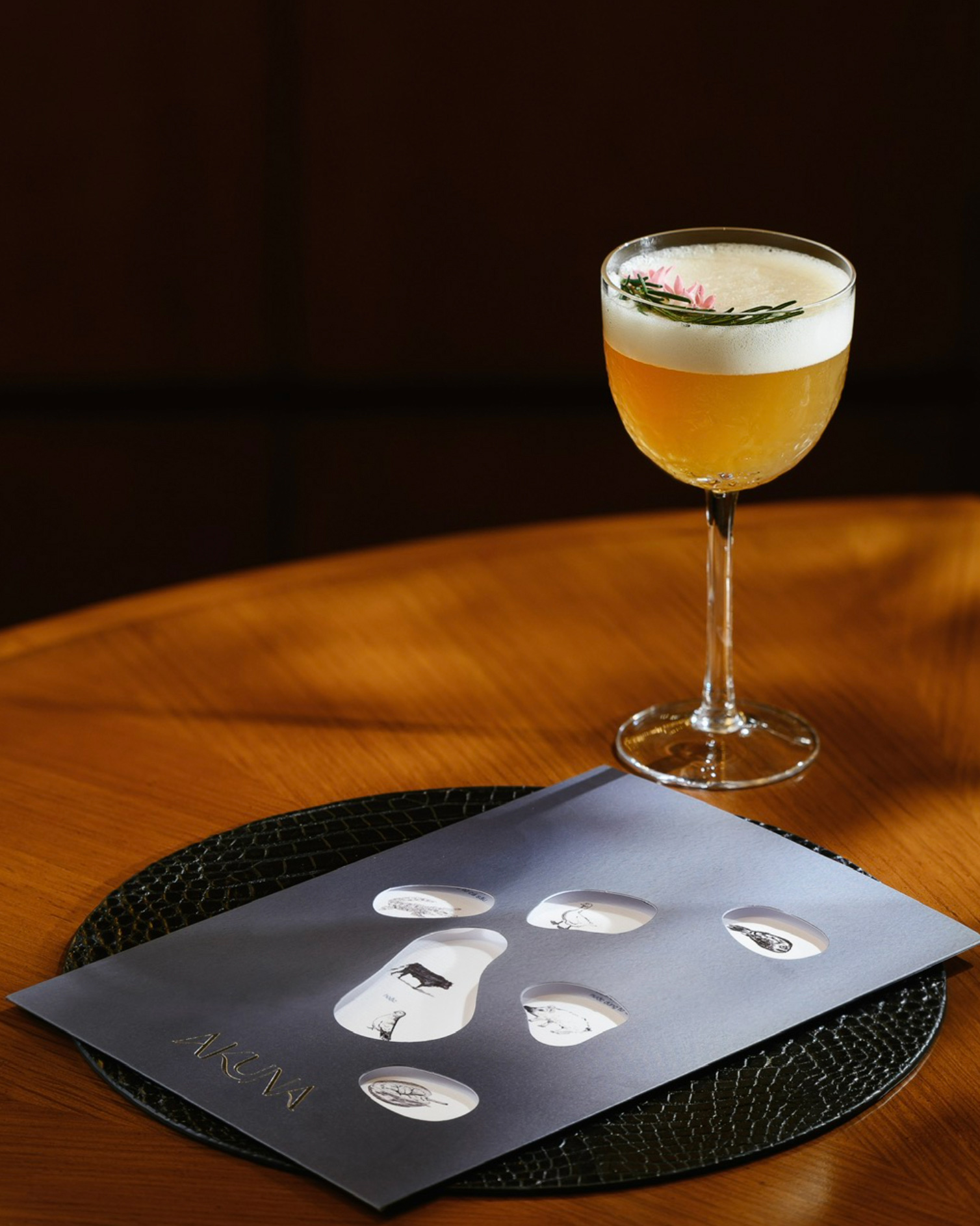 A glass of beer with frothy head sits on a wooden table, next to a sleek menu with abstract animal illustrations and the word 