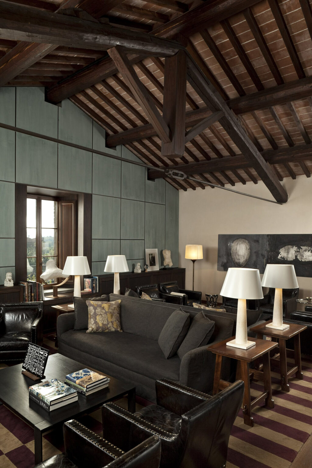 Chic living room with exposed beam ceiling, gray sofa, leather chairs, and table lamps. Art and sculptures on cabinets add elegance, warm ambiance.