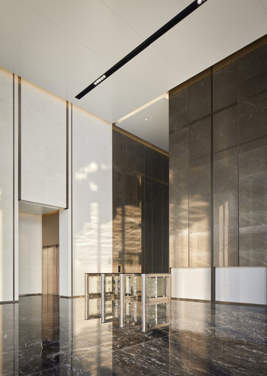Spacious modern office lobby with high ceilings, light and dark marble walls, reflective floor, and a sleek security gate. Soft natural light casts shadows. Calm ambiance.