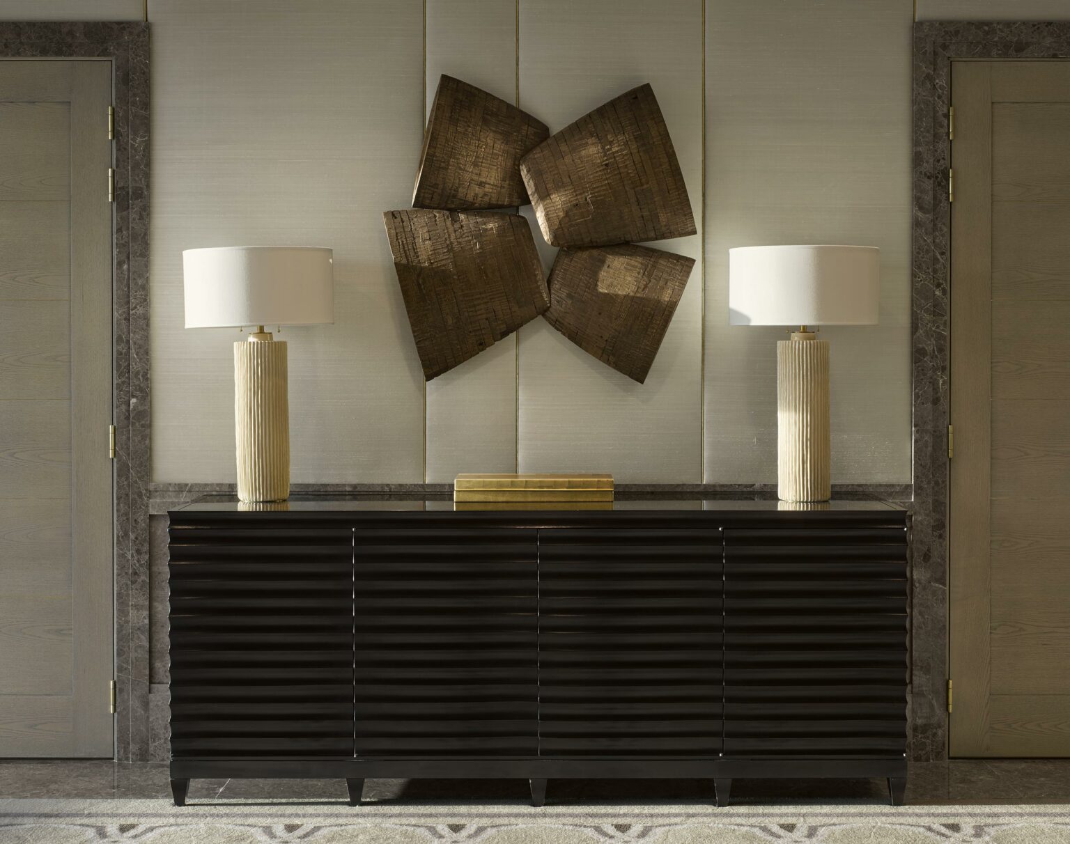 Elegant suite entrance with a textured dark console table, two beige lamps with white shades, and abstract wooden wall art, creating a sophisticated ambiance.