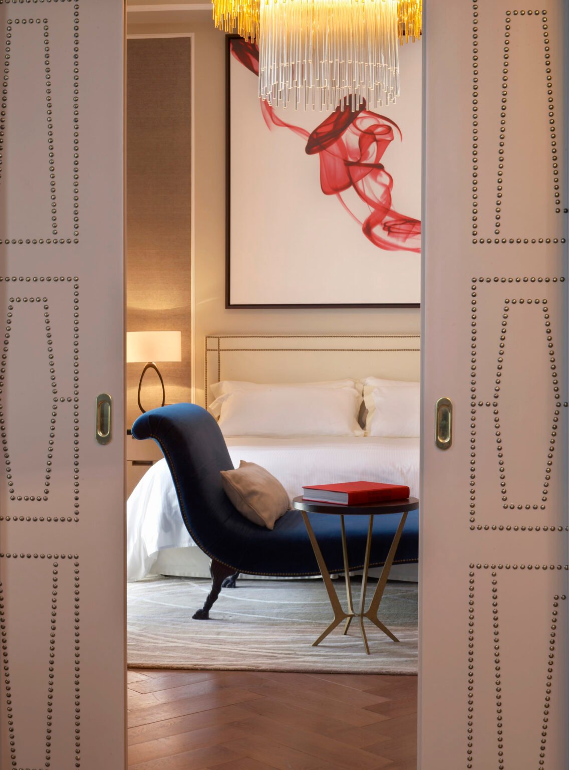 Elegant suite bedroom with a luxurious navy chaise lounge and beige pillow, beside an art deco gold side table with a red book. White bedding, modern art, and a chandelier add sophistication.