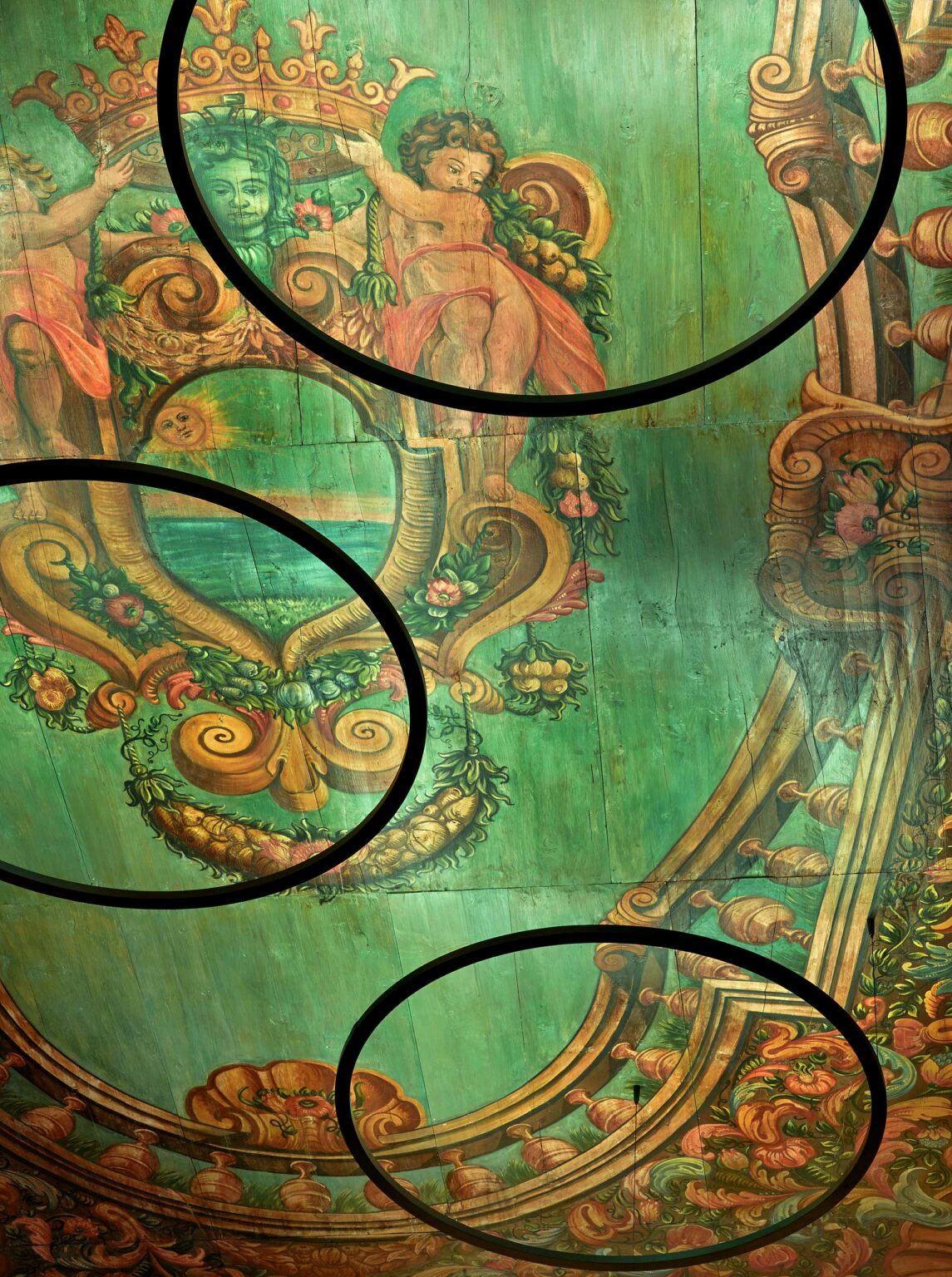 Hotel ceiling featuring a cherubic figure with rosy cheeks sitting amidst ornate Baroque scrolls and floral motifs on a vibrant green background, conveying a sense of historical elegance.