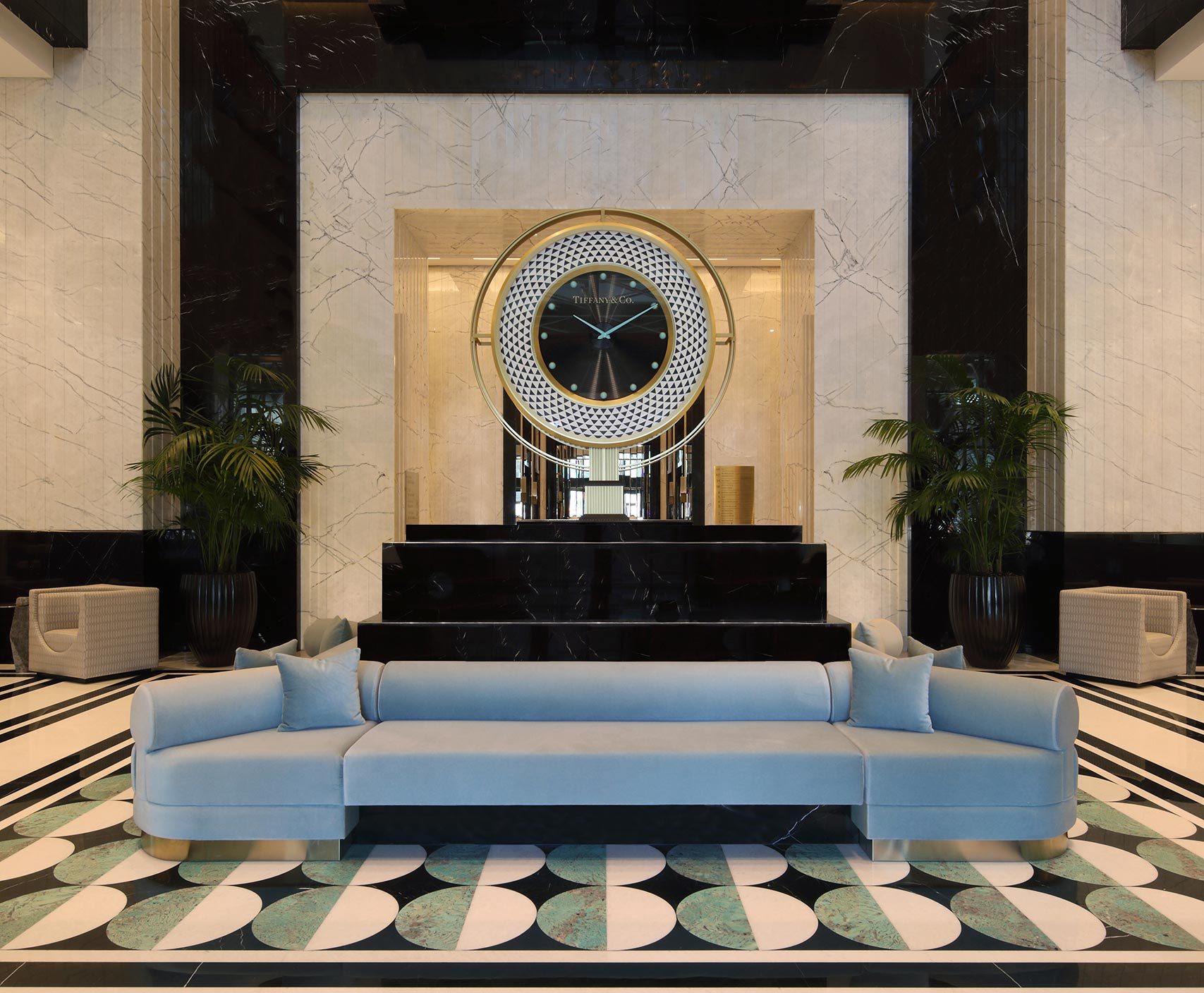 Luxurious hotel lobby with a large chandelier and a central decorative black and gold clock on a marble wall. A light blue sofa with pillows sits in front, flanked by tall green plants.