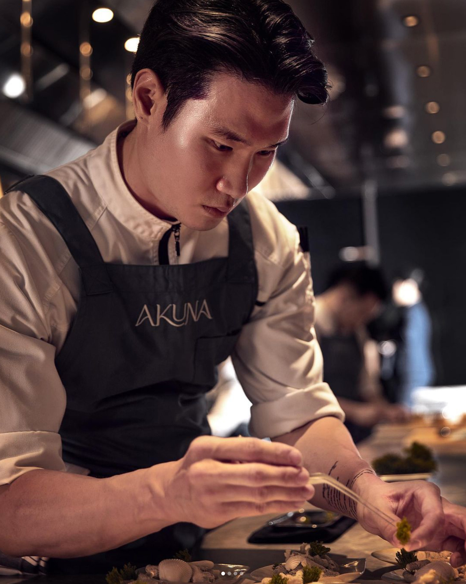 Chef in focus, wearing a white jacket and dark apron with the word 