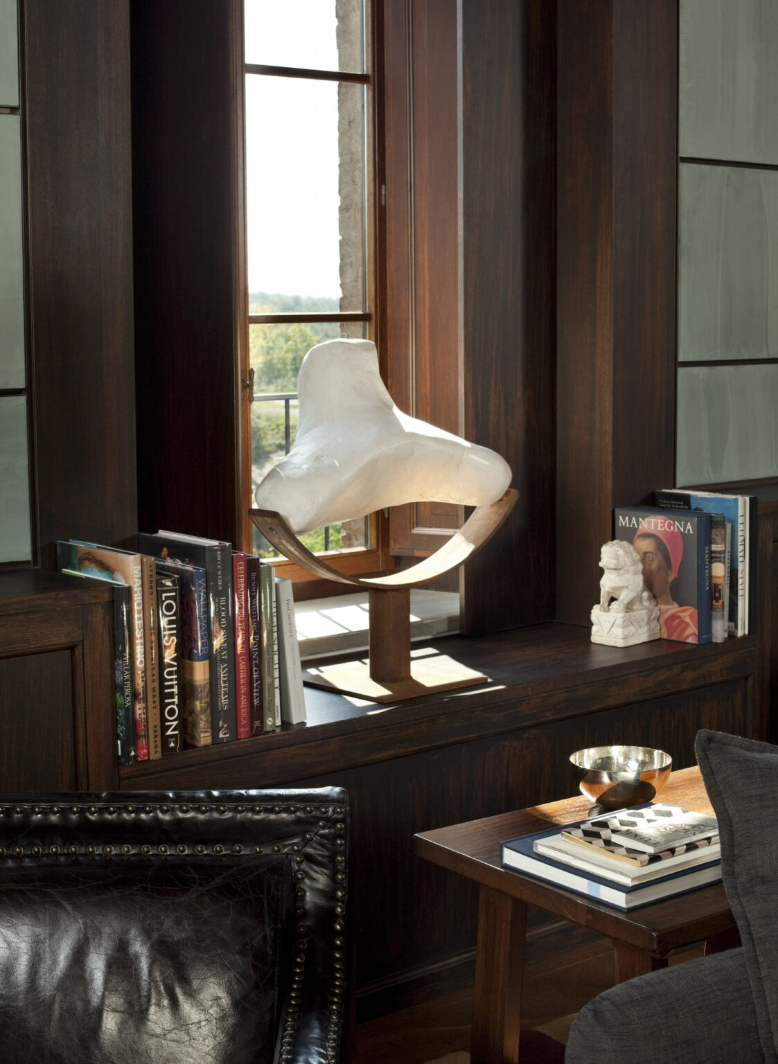 A white abstract sculpture sits on a wooden windowsill of a hotel lounge, surrounded by art books. Sunlight filters through a window, creating a serene, contemplative atmosphere.