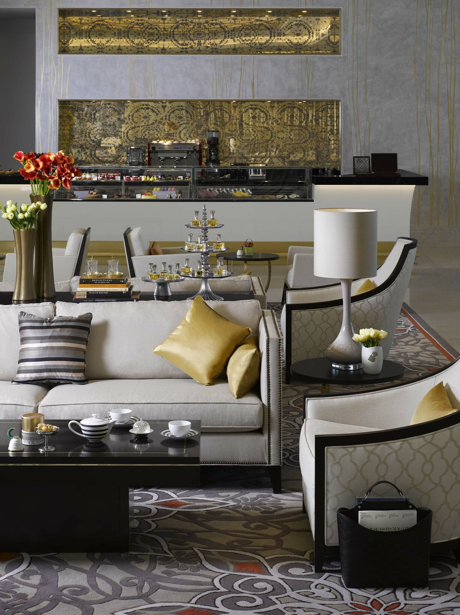 Elegant suite living room with plush beige sofas, gold pillows, and a coffee table set with teacups. A lavish dessert counter in the background.