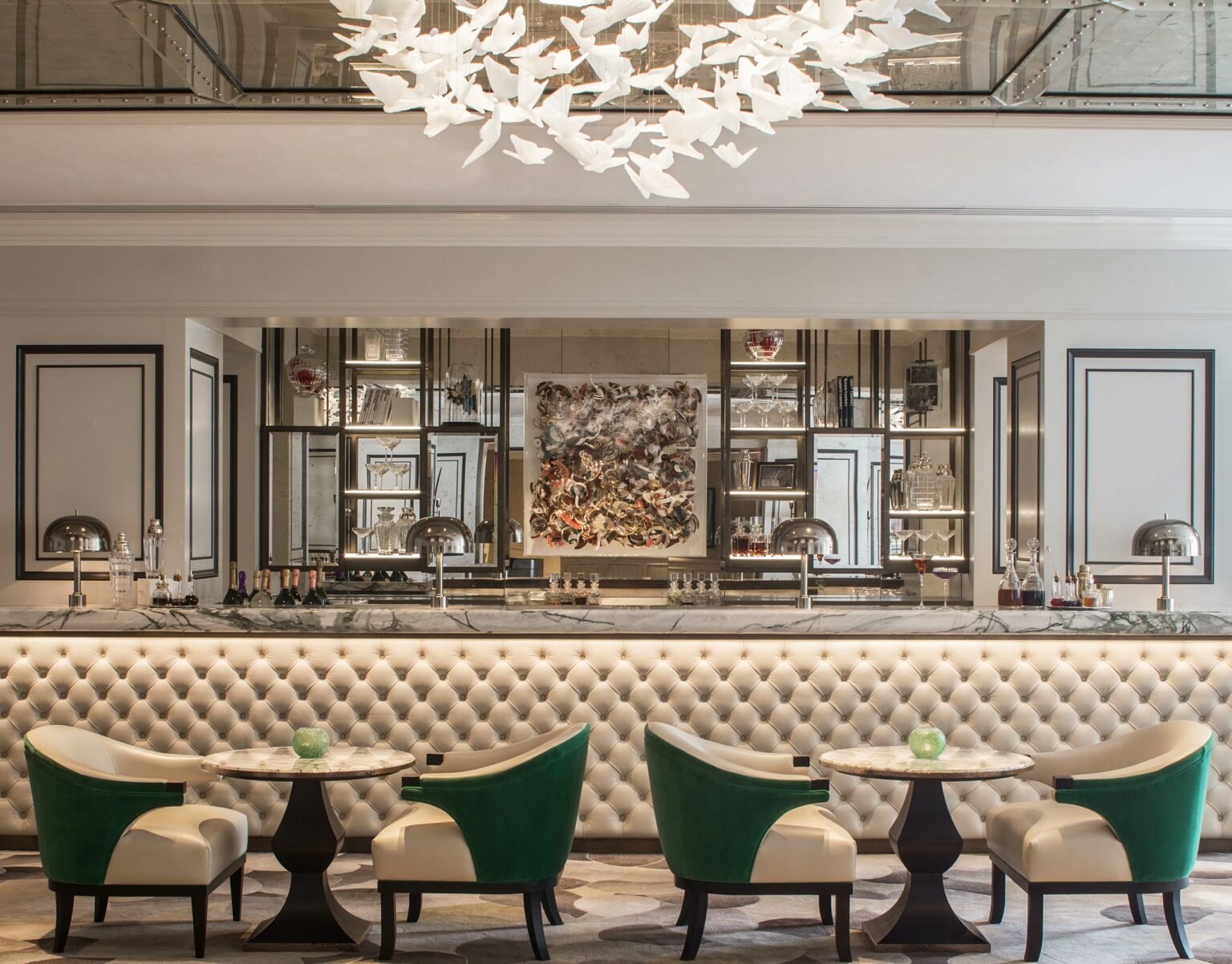 Elegant hotel bar interior with a tufted white counter, marble top, and metallic decor. Chic chairs with white and green accents surround tables. A modern chandelier exudes luxury.