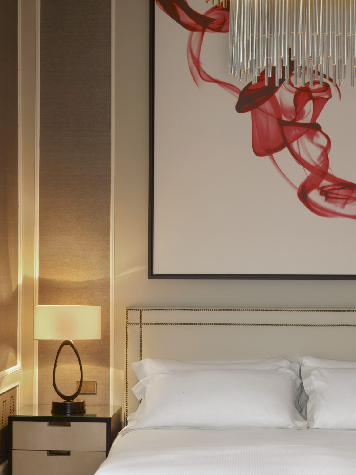 Elegant suite bedroom with soft lighting. A modern lamp sits on a nightstand next to a bed with white linens. Above, abstract art with flowing red shapes adds vibrancy.
