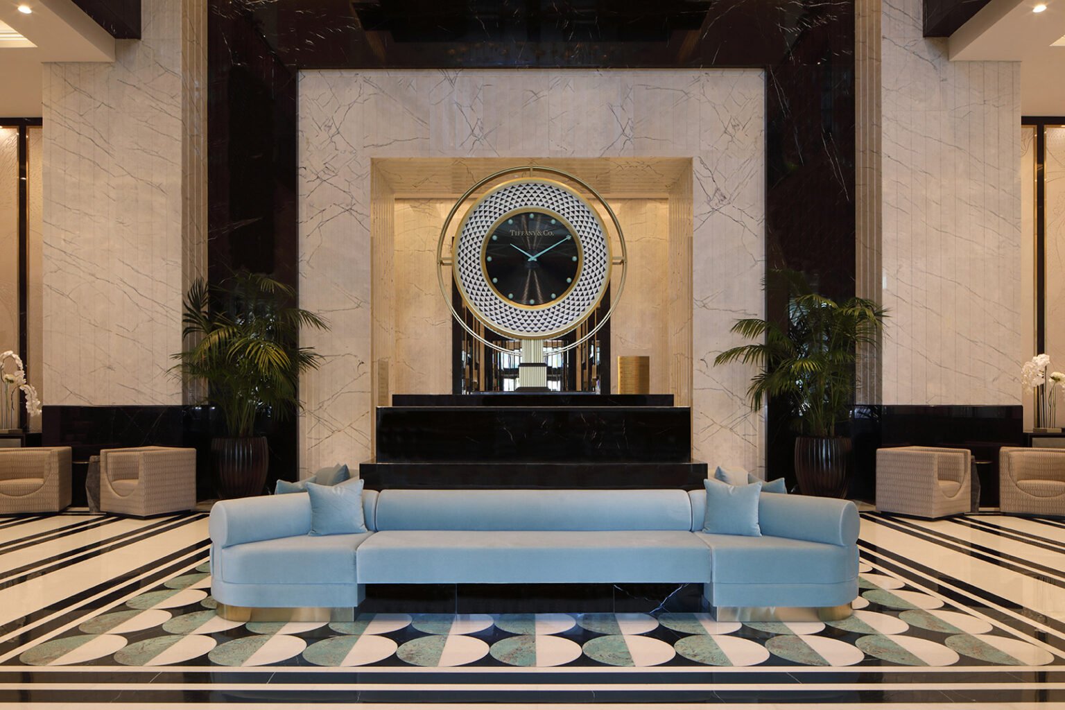 Luxurious hotel lobby with a large black and gold clock on a marble wall. A light blue sofa with pillows sits in front, flanked by tall green plants.