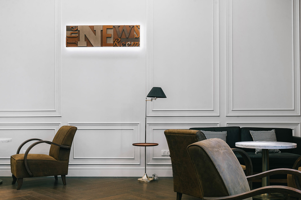 Cozy lounge with brown armchairs, a floor lamp, and a small table against a paneled white wall. A lit sign reads 