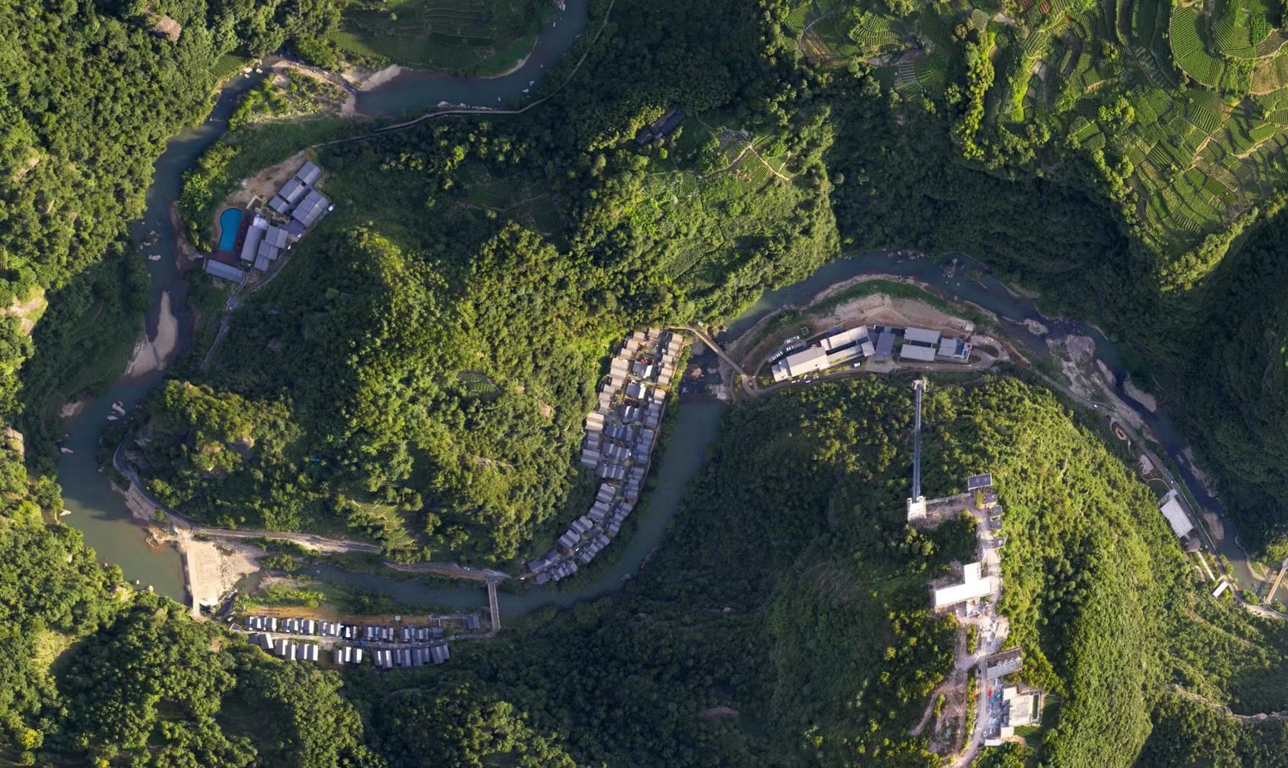 Aerial view of lush green hills with a winding river and scattered buildings. The vibrant landscape conveys a peaceful, natural setting.