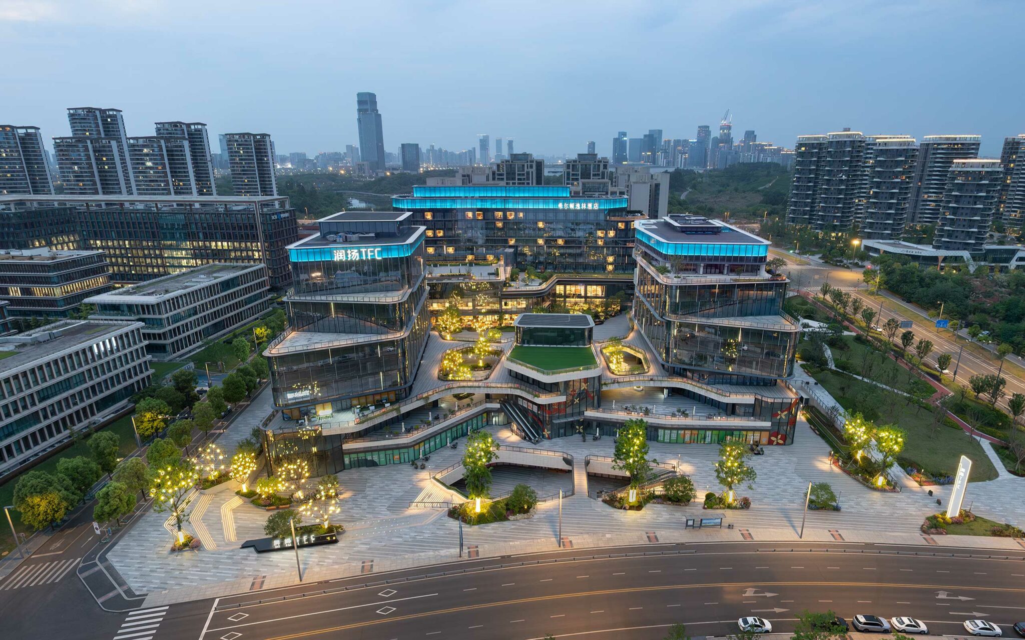 Aerial view of a modern, illuminated commercial complex at dusk. Buildings have blue accents and are surrounded by greenery and pathways. City skyline in the background.