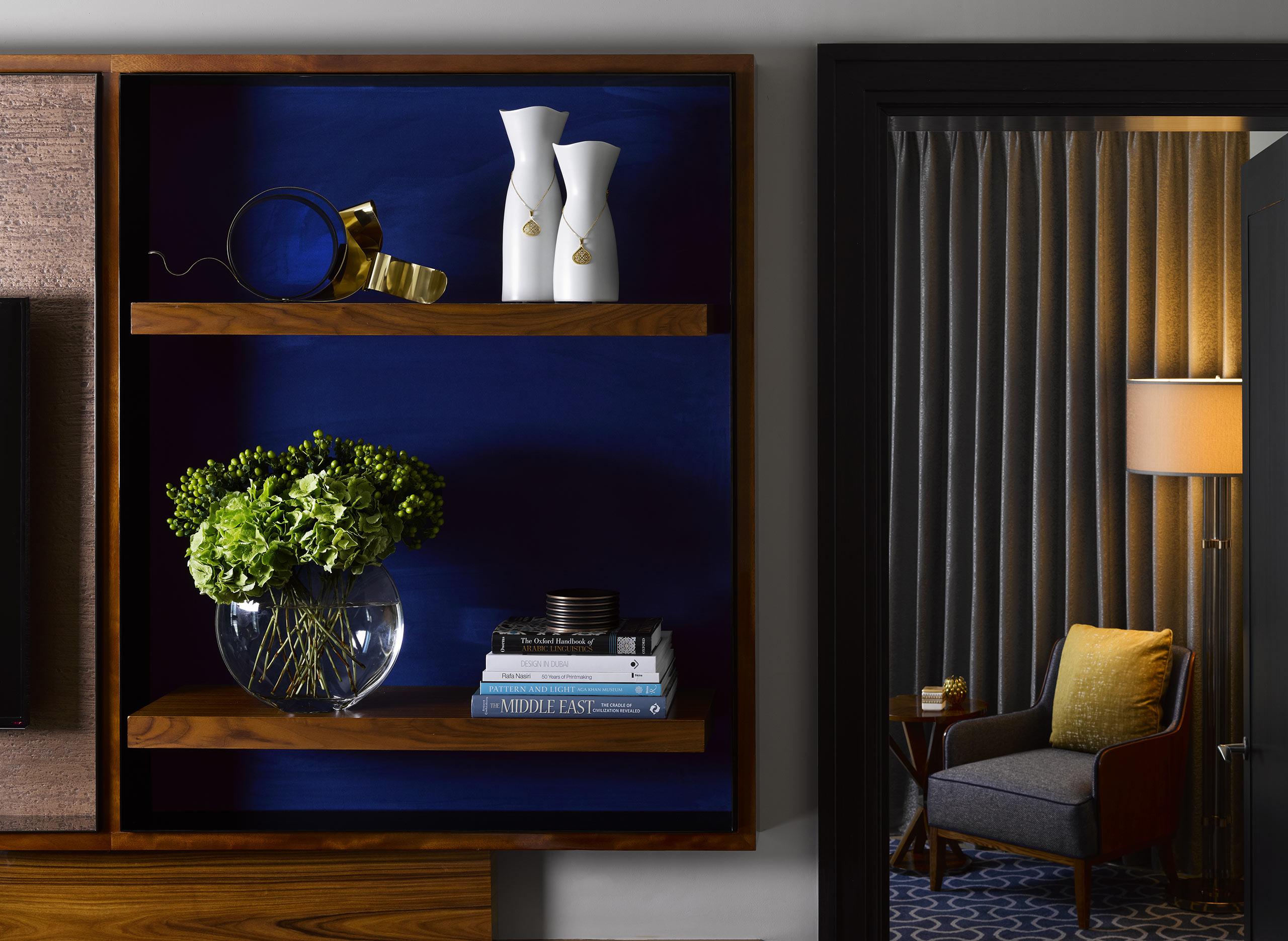 A modern wooden shelf against a blue wall holds decorative white vases and golden rings on top, with green hydrangeas and books below. A cozy suite with a lamp and chair is partially visible on the right. The tone is elegant and serene.