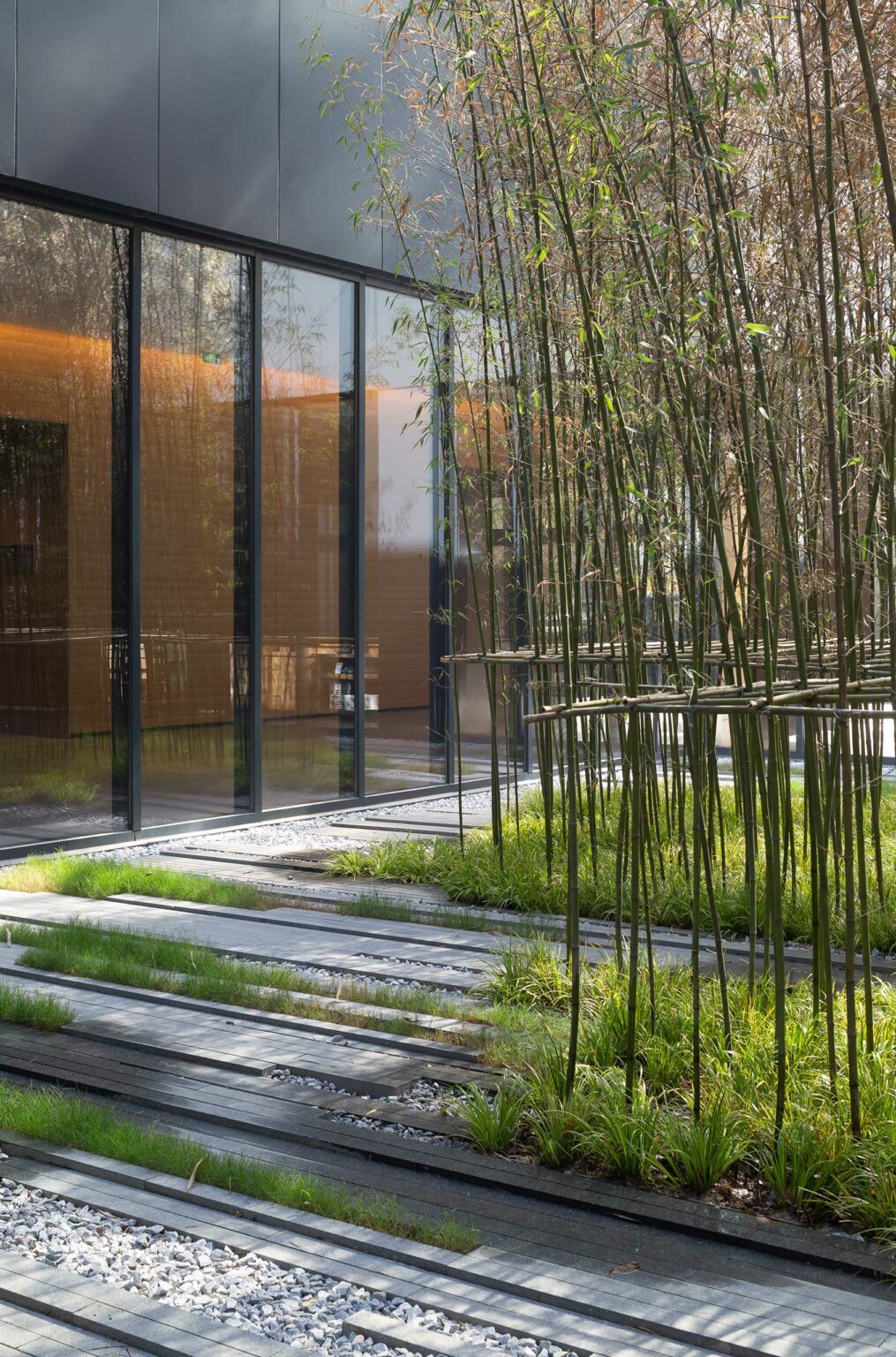 Modern hotel architecture with floor-to-ceiling windows reflecting bamboo garden. Pathway with grass and pebbles creates a serene, natural ambiance.