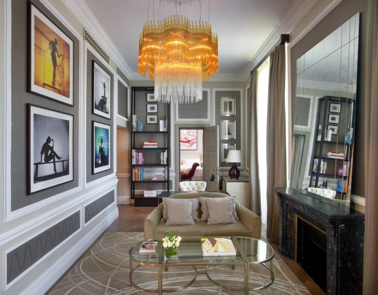 Elegant suite living room with modern decor, featuring a beige sofa, large chandelier, framed silhouette art, and a mirrored fireplace. Bookshelves add a cozy touch.