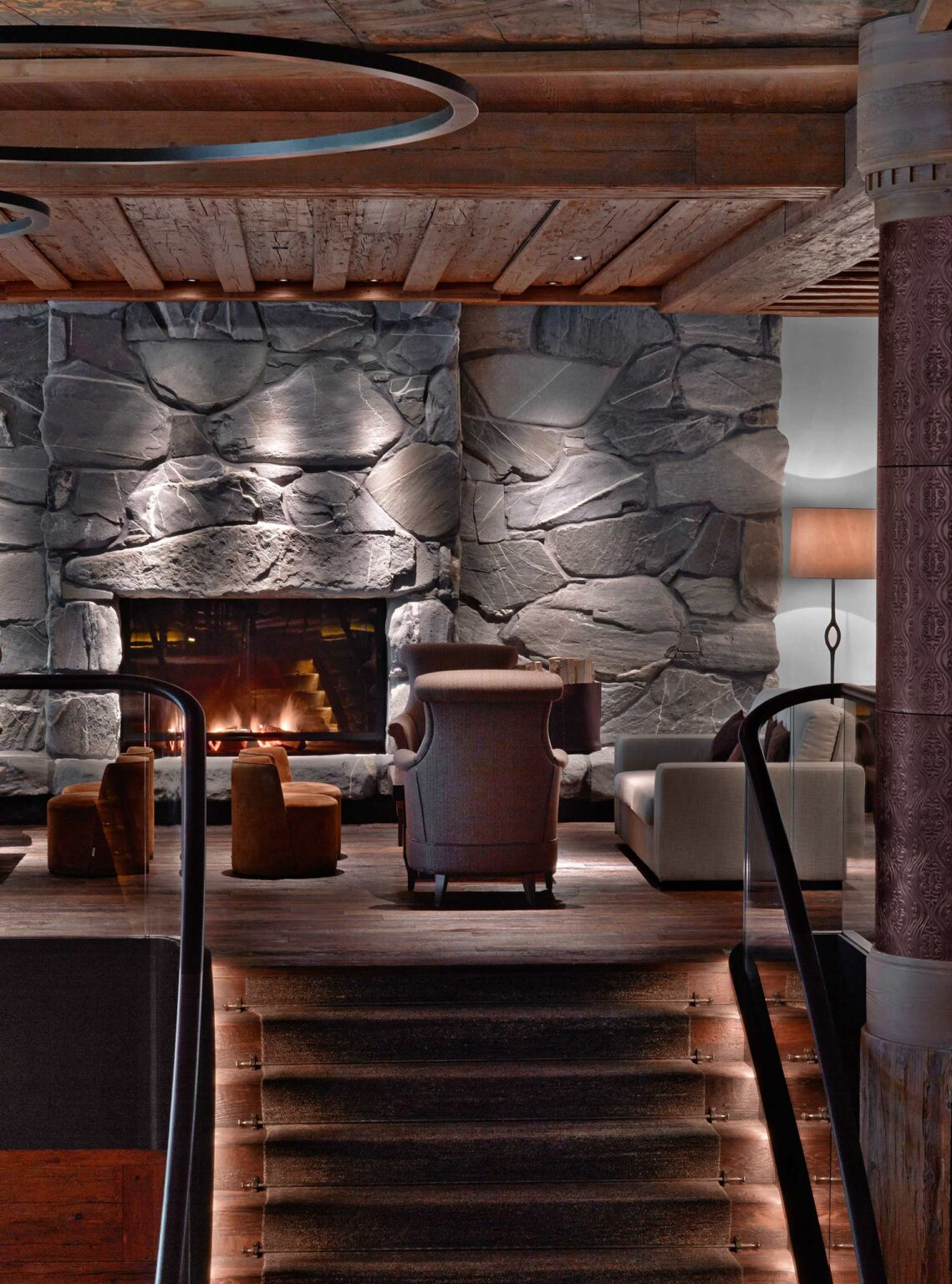 Cozy hotel lounge with stone fireplace, warm fire, and plush armchairs. Soft lighting enhances the rustic wood ceiling, creating a relaxed and inviting ambiance.