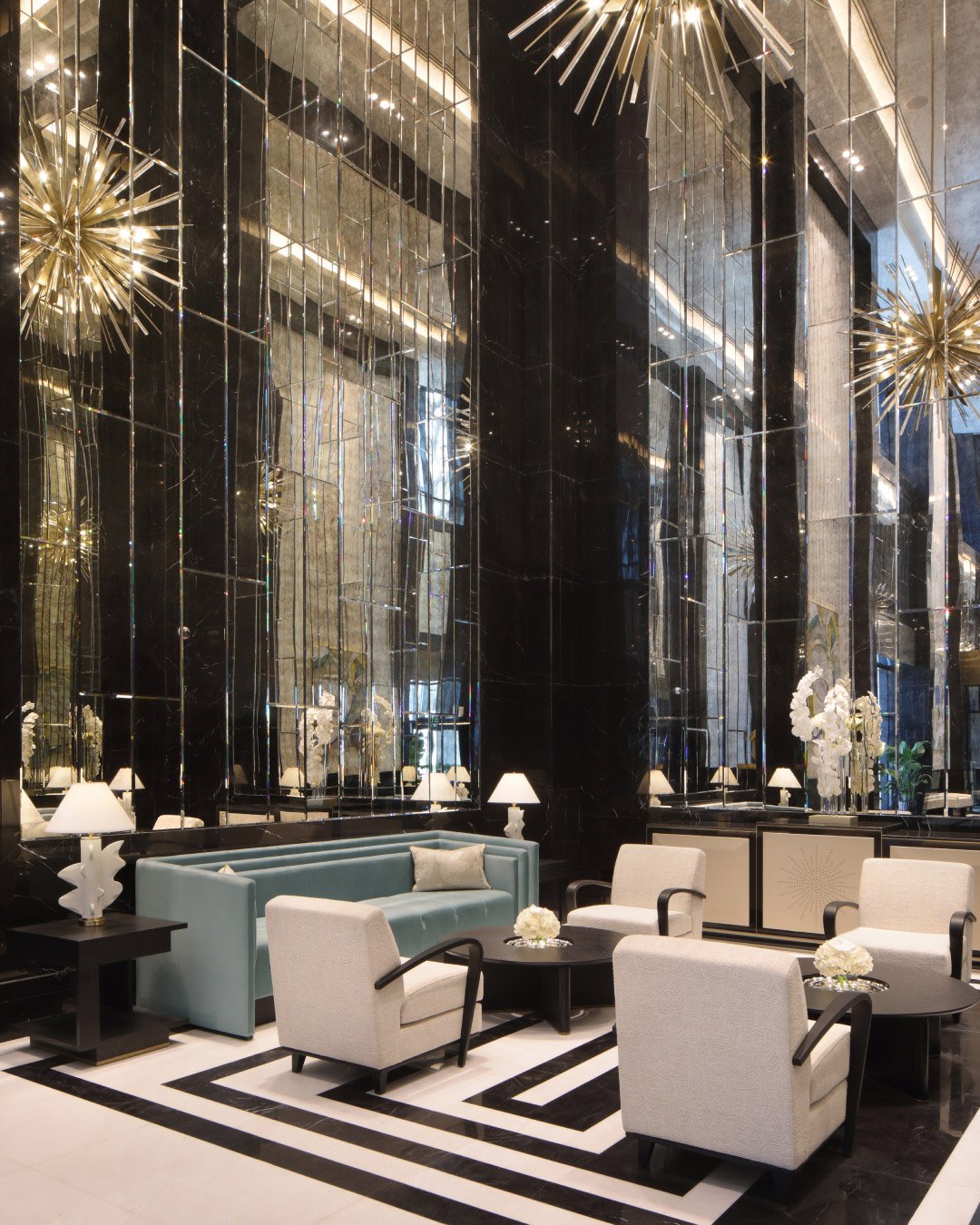 Luxurious hotel lobby with tall black mirrored walls, a light blue sofa, beige chairs, and starburst chandeliers. The tone is elegant and sophisticated.