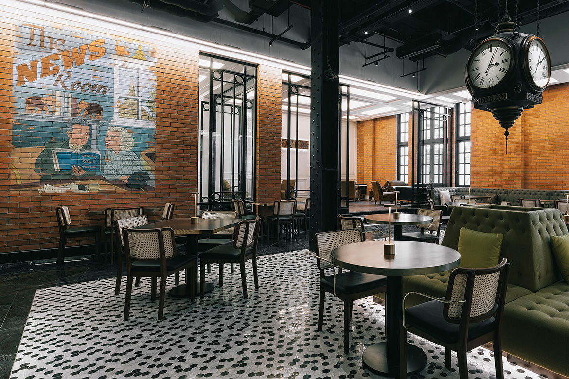 Spacious hotel cafe with brick walls, vintage mural of two people reading, and a large clock. Cozy seating, natural light, and a relaxed atmosphere.
