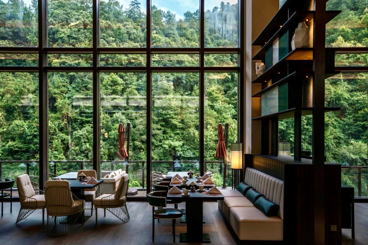 Luxurious hotel dining area with large windows offering a lush forest view. Elegant tables, plush chairs, and a serene, sophisticated ambiance.