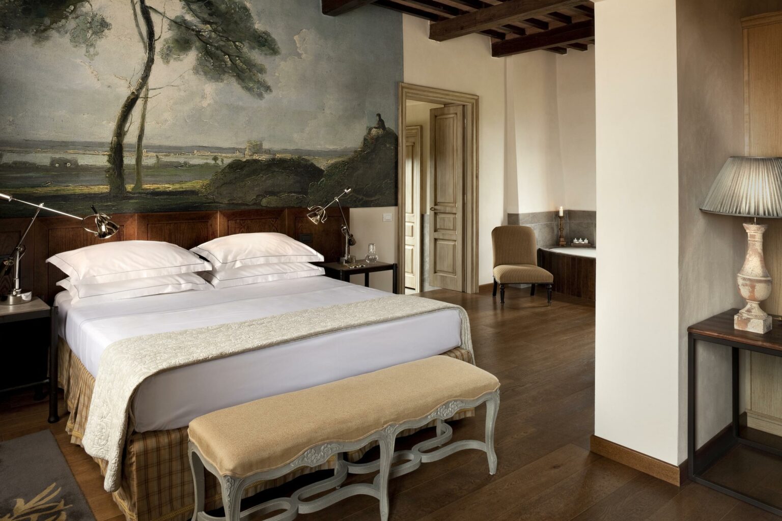 Elegant hotel bedroom with a king-size bed, rustic wooden decor, and a serene wall mural of a landscape. Warm lighting and cozy seating create a peaceful ambiance.