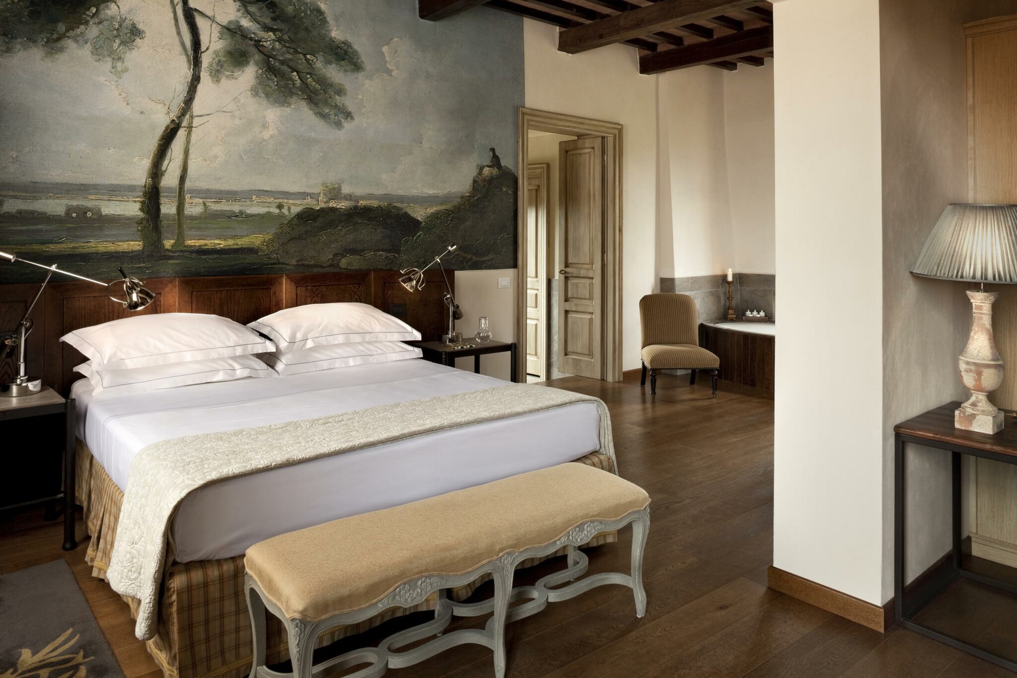Elegant hotel bedroom with a king-size bed, rustic wooden decor, and a serene wall mural of a landscape. Warm lighting and cozy seating create a peaceful ambiance.