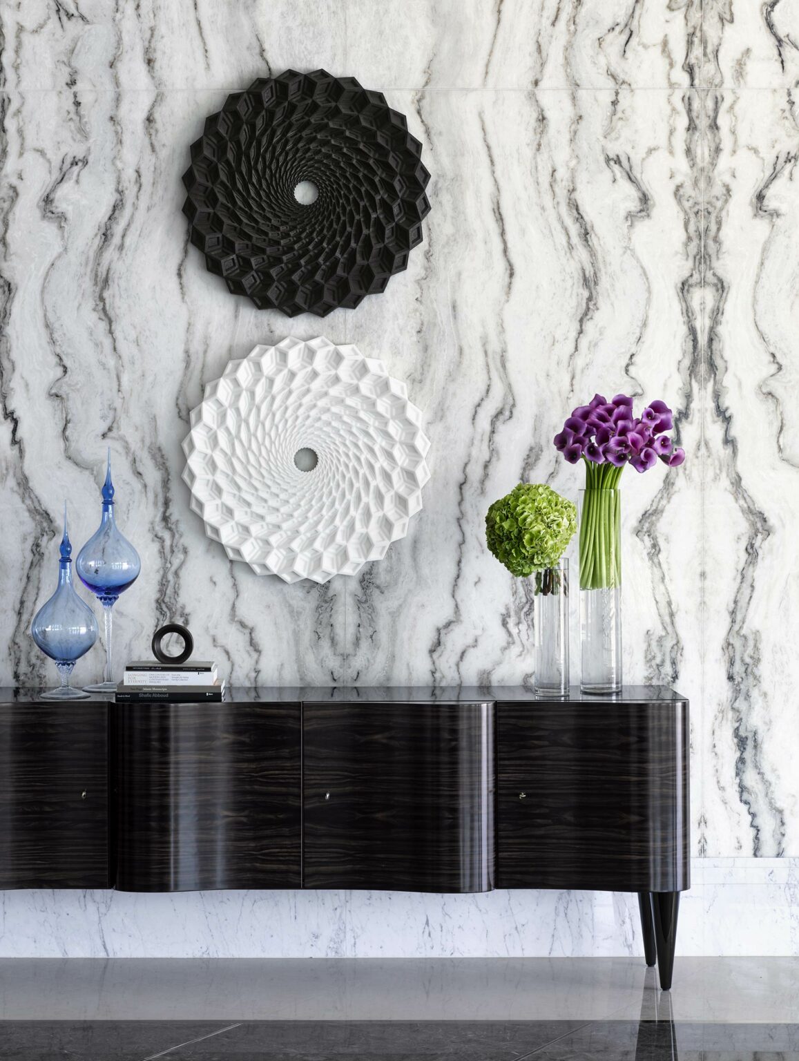 Modern hotel interior with two circular, geometric wall art pieces, one black and one white, on a marbled wall. Below, a sleek dark console holds glass vases with purple and green flowers, adding elegance.