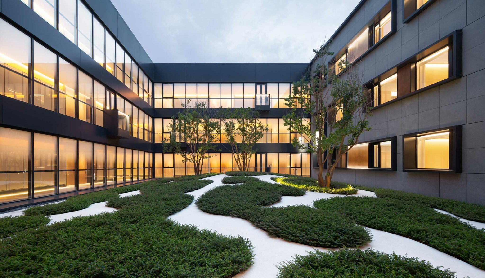Modern hotel building courtyard with geometric shrub patterns on white gravel. Warmly lit windows line the three-story structure, creating a serene evening ambiance.