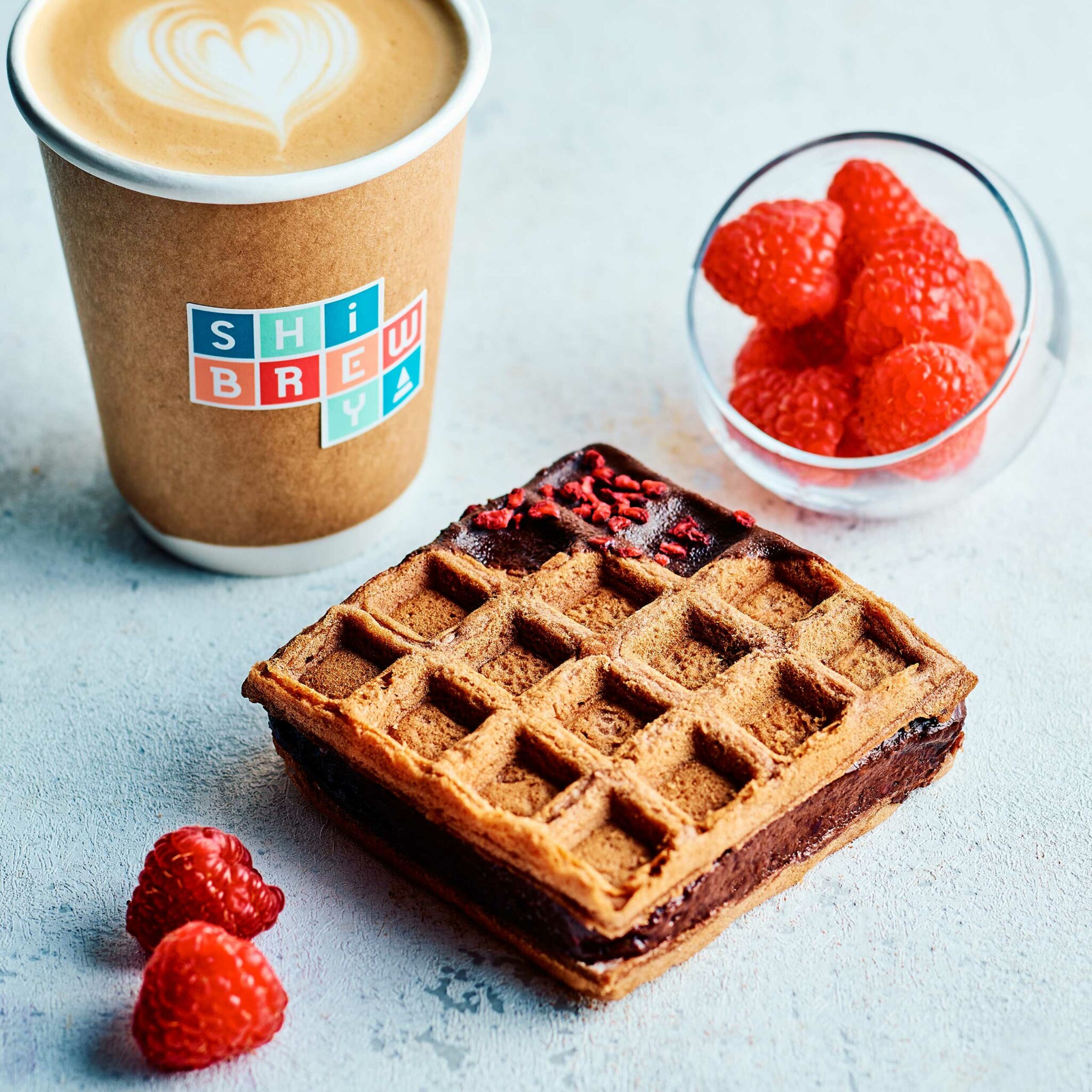 A latte with heart-shaped foam in a branded cup, beside a chocolate-filled waffle and fresh raspberries, conveys a cozy, indulgent breakfast mood.