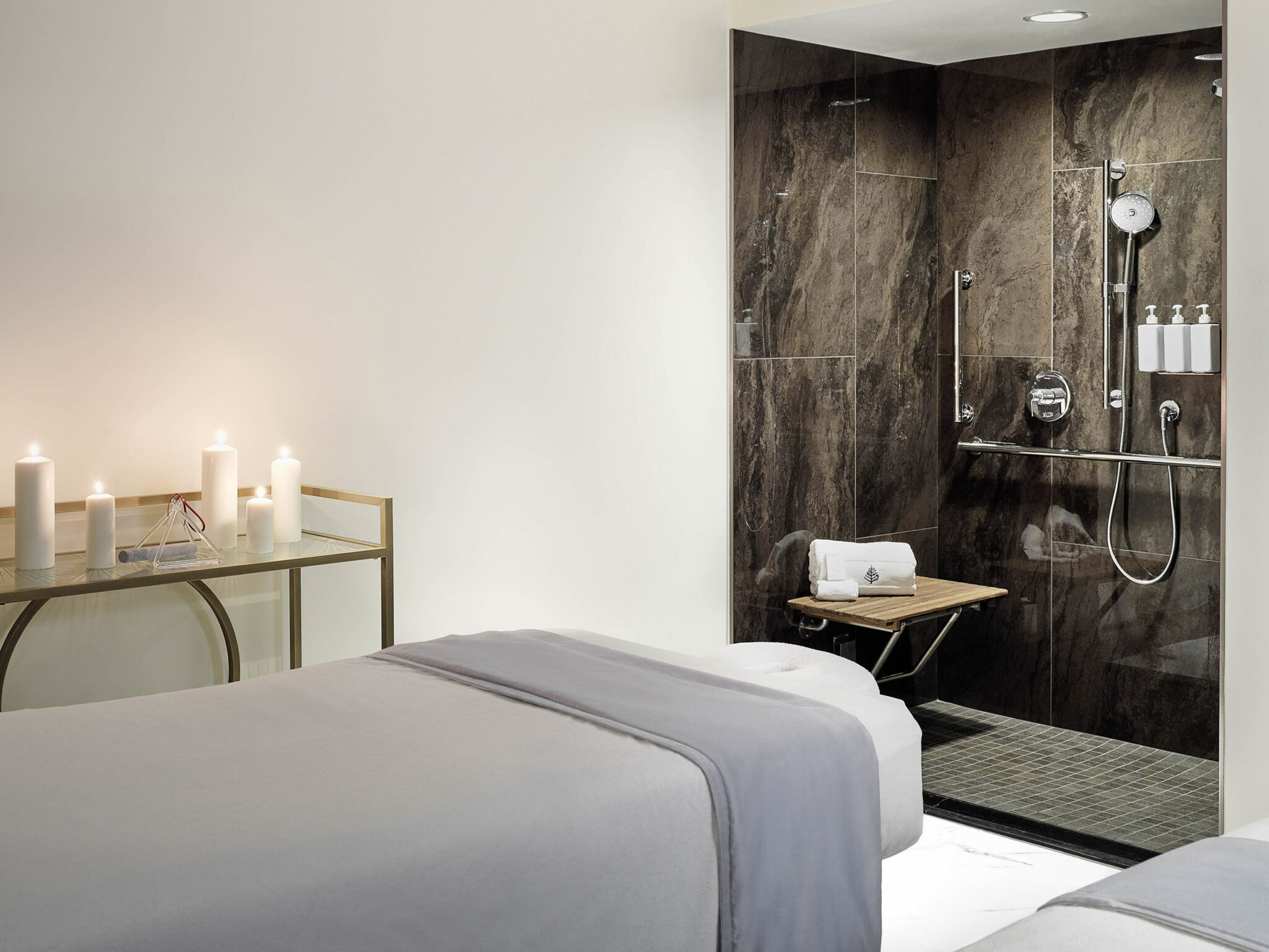 A serene hotel spa treatment room with a massage table draped in white and gray sheets, glowing candles on a glass table, and a modern rain shower in the corner.