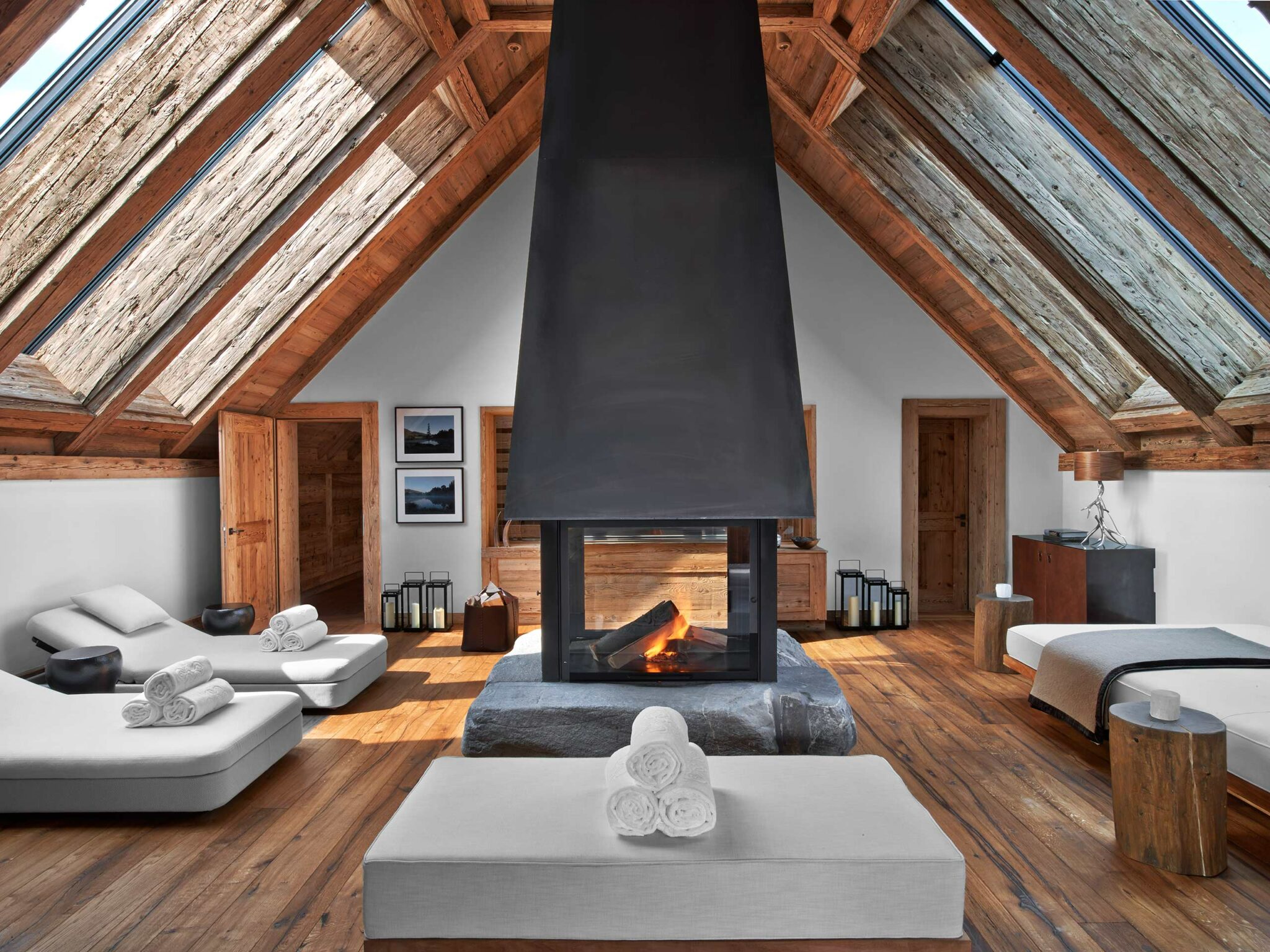 Luxurious hotel spa room with a central modern fireplace, wooden beams, and skylights. White loungers with rolled towels create a serene, inviting atmosphere.