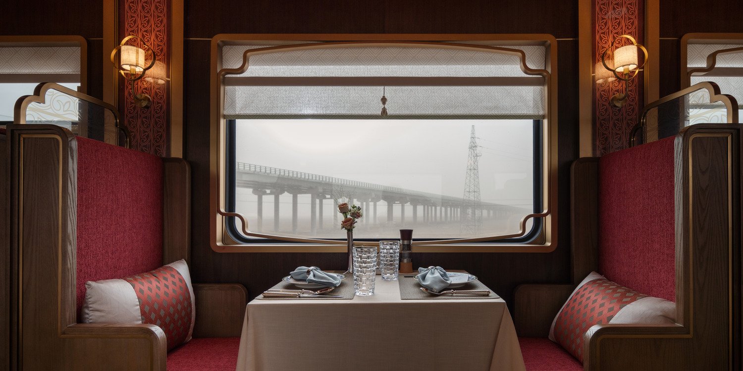 Luxurious train interior with elegant wooden paneling and large windows showing mountain views. Plush seating with decorative pillows creates a cozy atmosphere.