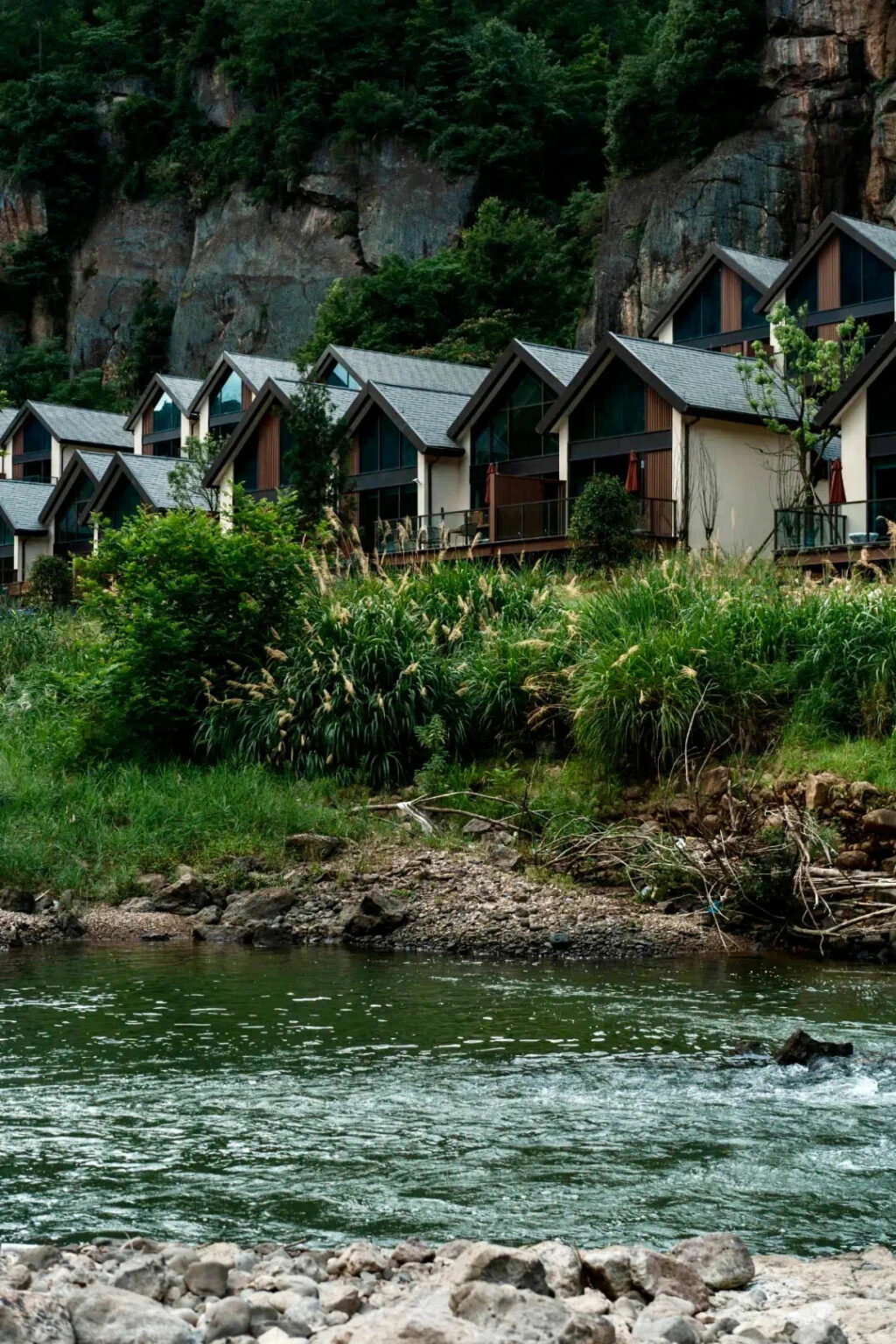 Modern cabins with A-frame roofs are nestled against a lush green hillside, overlooking a gentle river. The scene conveys tranquility and natural beauty.