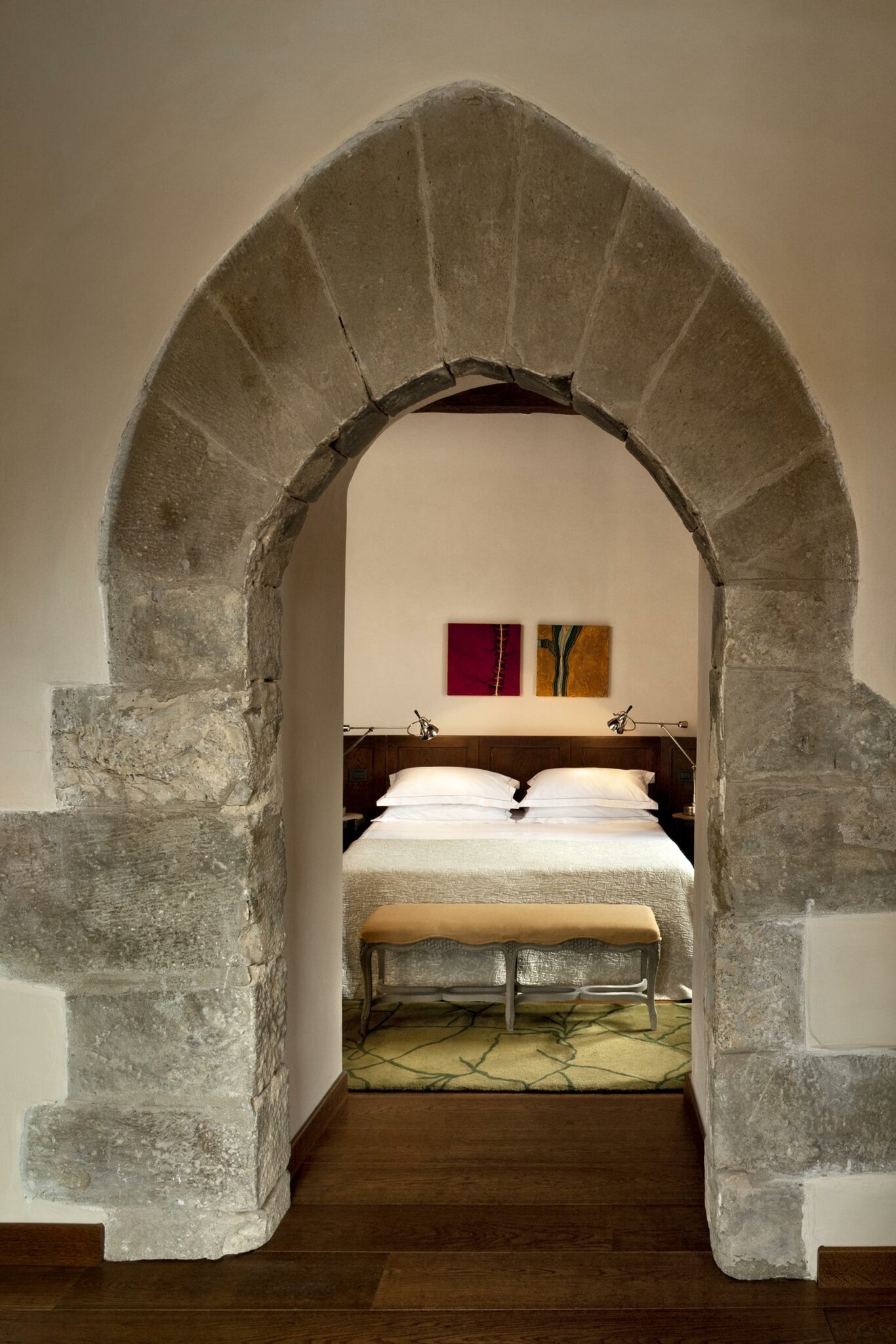 Stone archway leads to a cozy hotel bedroom with a neatly made bed, wooden headboard, and wall art. Warm tones create a welcoming, serene atmosphere.