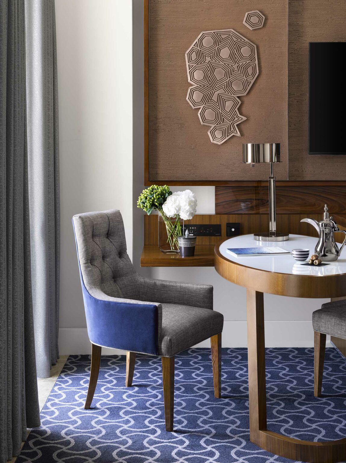 Modern hotel guest room corner featuring a grey and blue upholstered chair next to a round wooden table. Wall art and a vase of flowers add elegance.