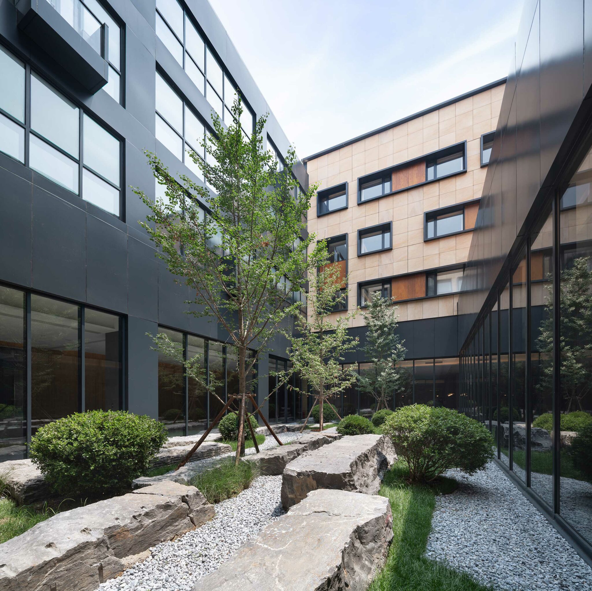 Modern hotel courtyard with tall buildings, large rocks, greenery, and a gravel path. The scene is serene and well-maintained, highlighting urban nature.