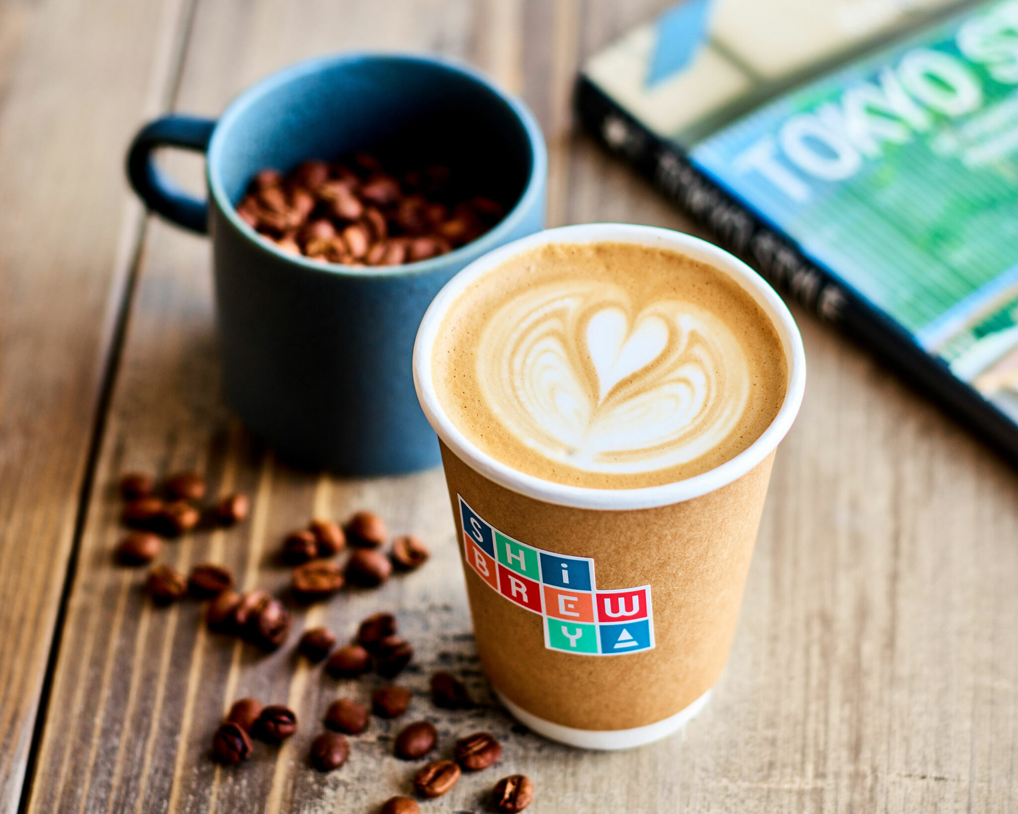 Latte with heart latte art in paper cup on wooden table, next to scattered coffee beans and a blue mug filled with beans, creating a cozy ambiance.