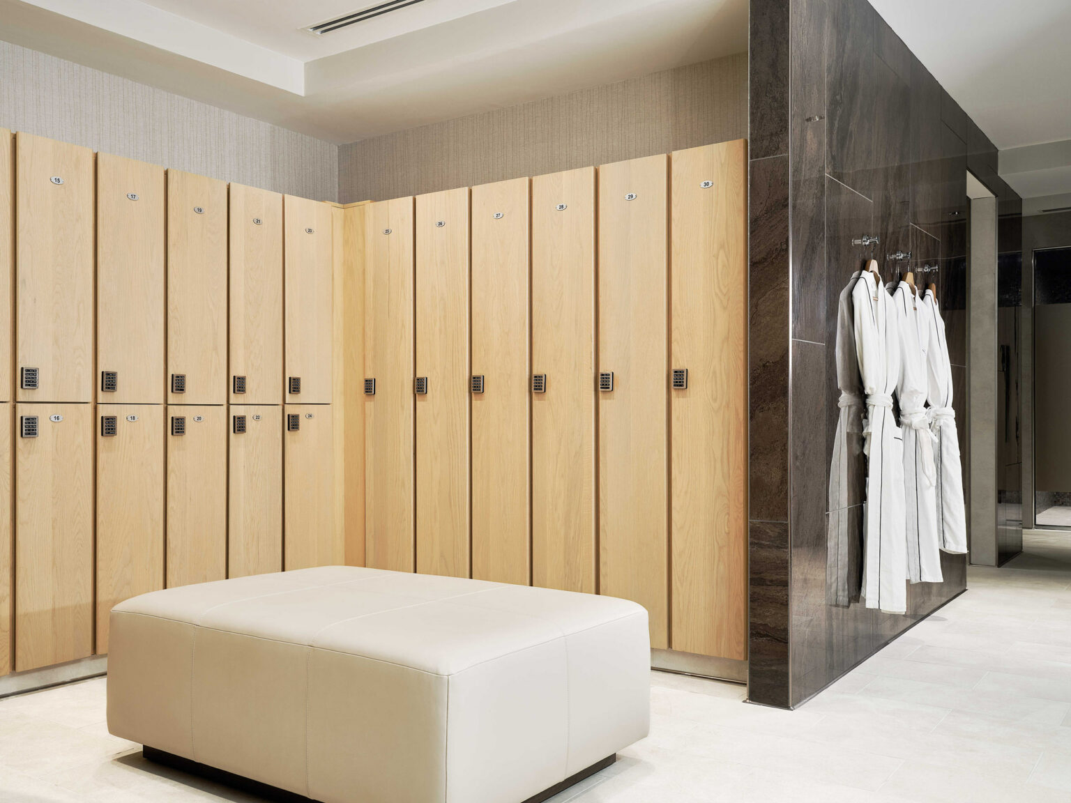Modern hotel spa locker room with light wood lockers, digital locks, beige cushioned bench, and wall with white bathrobes, conveying cleanliness and order.