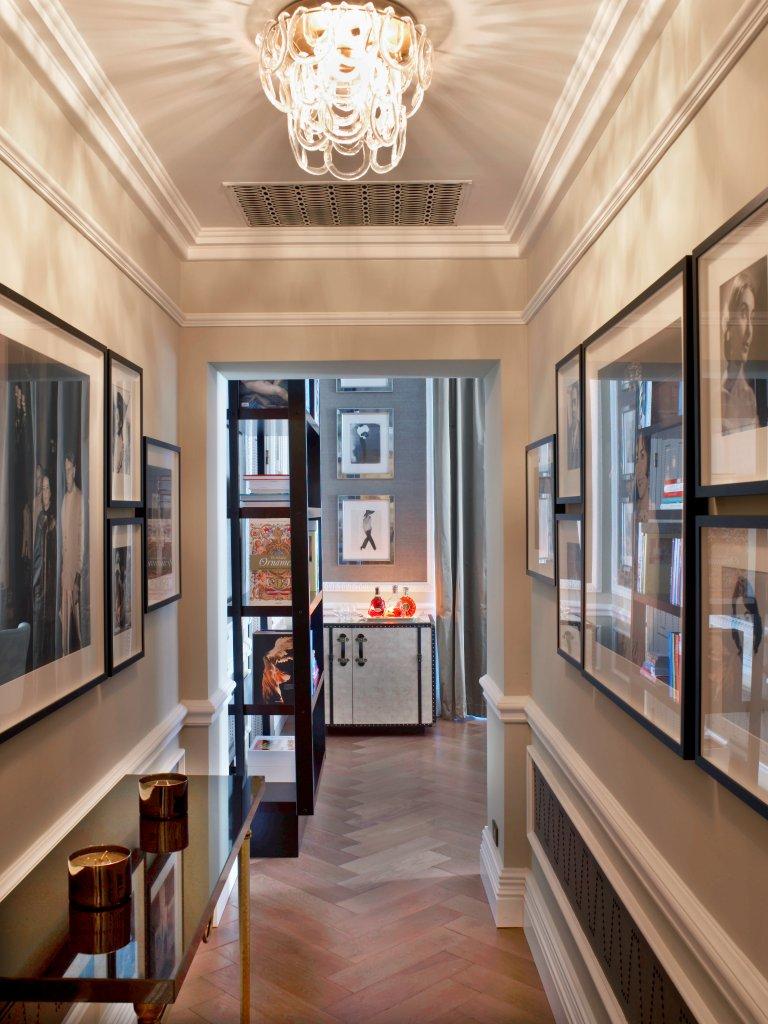 Elegant hotel hallway with framed photos, sleek black table, and warm chandelier lighting. Leads to a cozy room with shelves and decorative items, exuding sophistication.