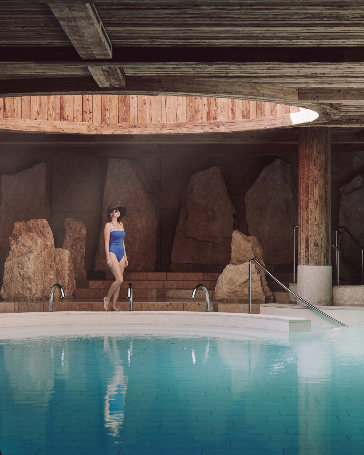 A woman in a blue swimsuit and wide-brimmed hat stands by an indoor hotel pool with rocky walls. The warm lighting creates a serene, luxurious vibe.