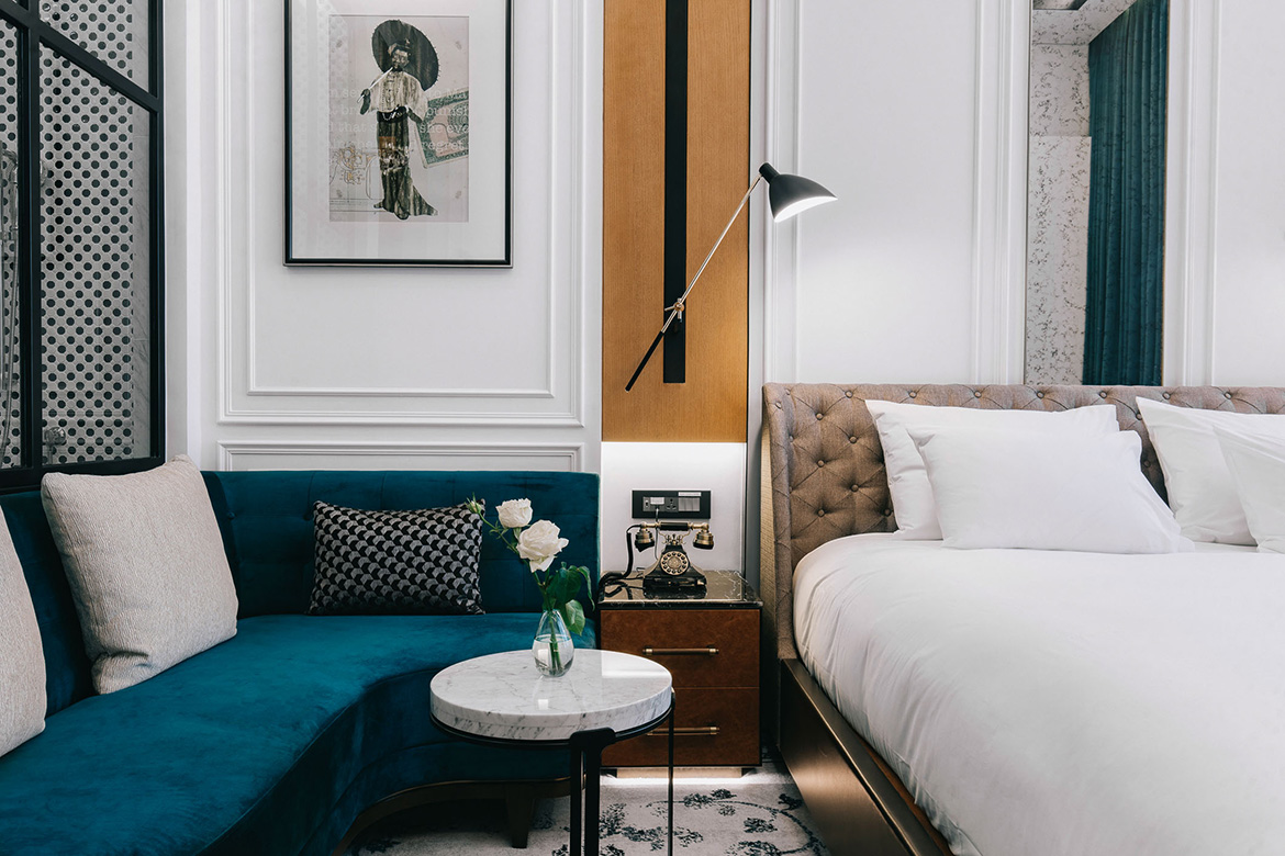 Stylish hotel room with a tufted bed, blue velvet sofa, marble table with flowers, and a vintage phone. Elegant artwork and warm lighting create a cozy feel.