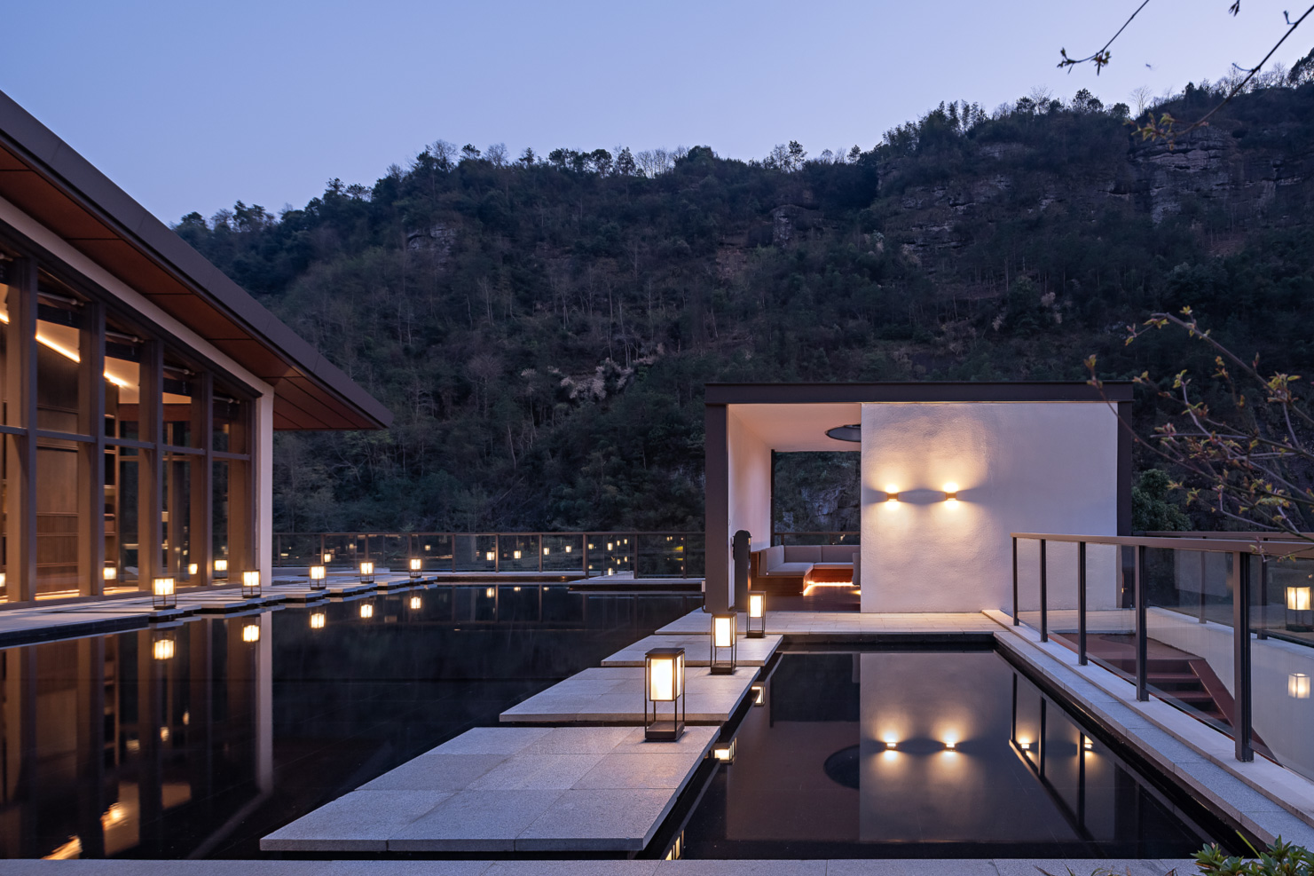 Rooftop terrace at dusk with soft lighting, framed by a modern building on the left and a cozy seating area on the right. Surrounded by a forested mountain backdrop.