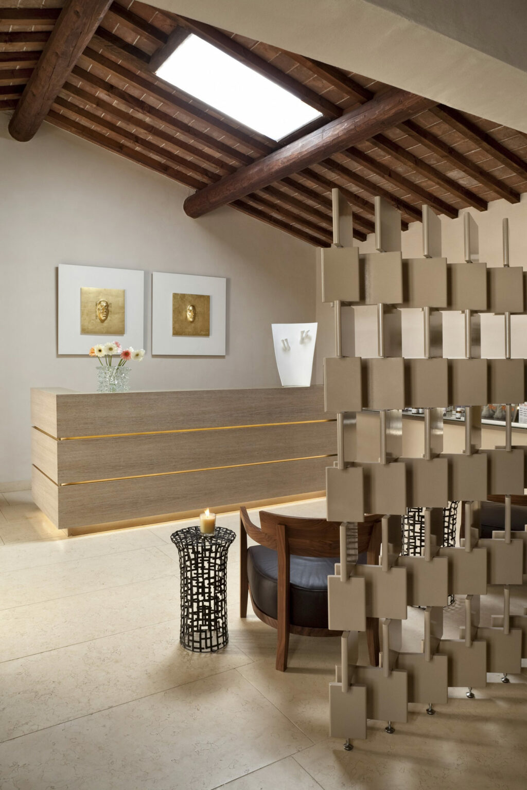 Modern spa reception with a wooden ceiling and geometric divider. Features a minimalist desk, wall art, a vase with flowers, and a lit candle on a metal stand.