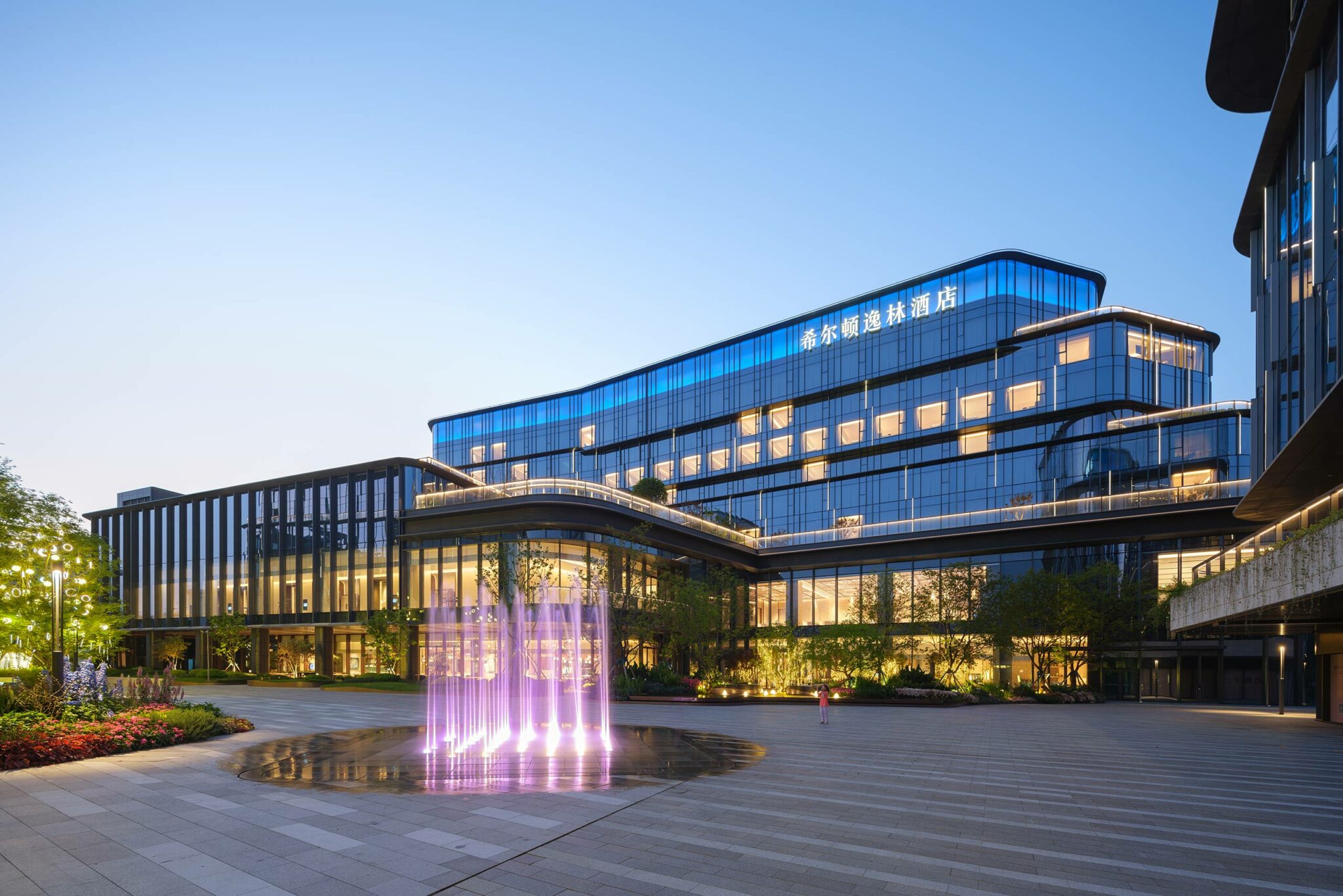 Modern hotel exterior, showing a glass building at dusk with illuminated windows and a vibrant purple fountain in front. The scene feels tranquil and contemporary.
