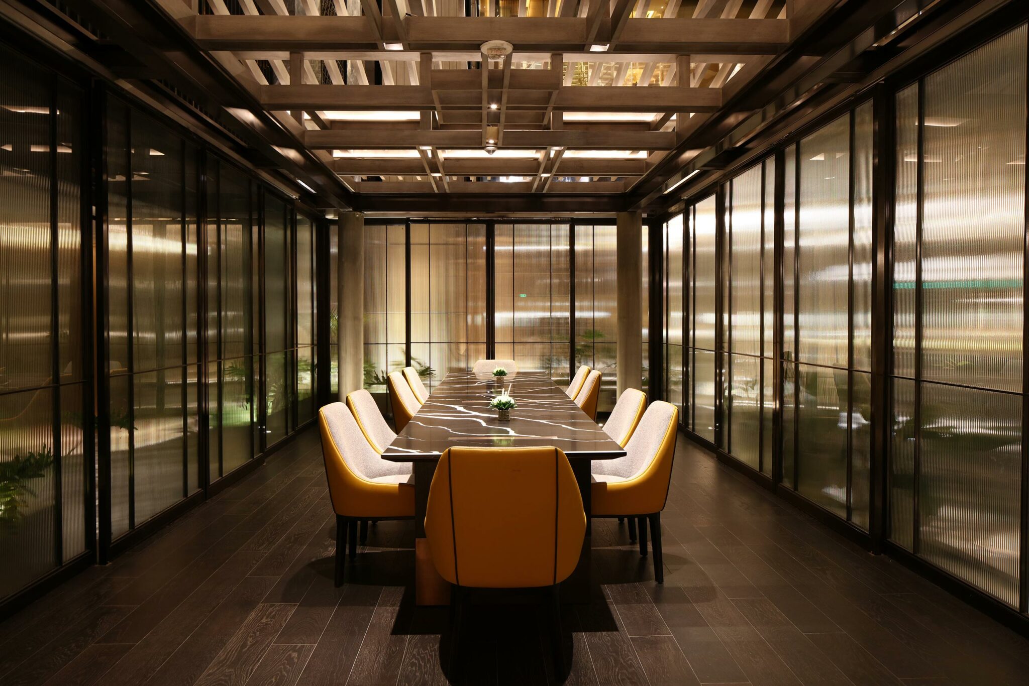 Sleek private dining room with a long marble table, surrounded by modern yellow chairs. Glass walls and dim lighting create an elegant, sophisticated atmosphere.