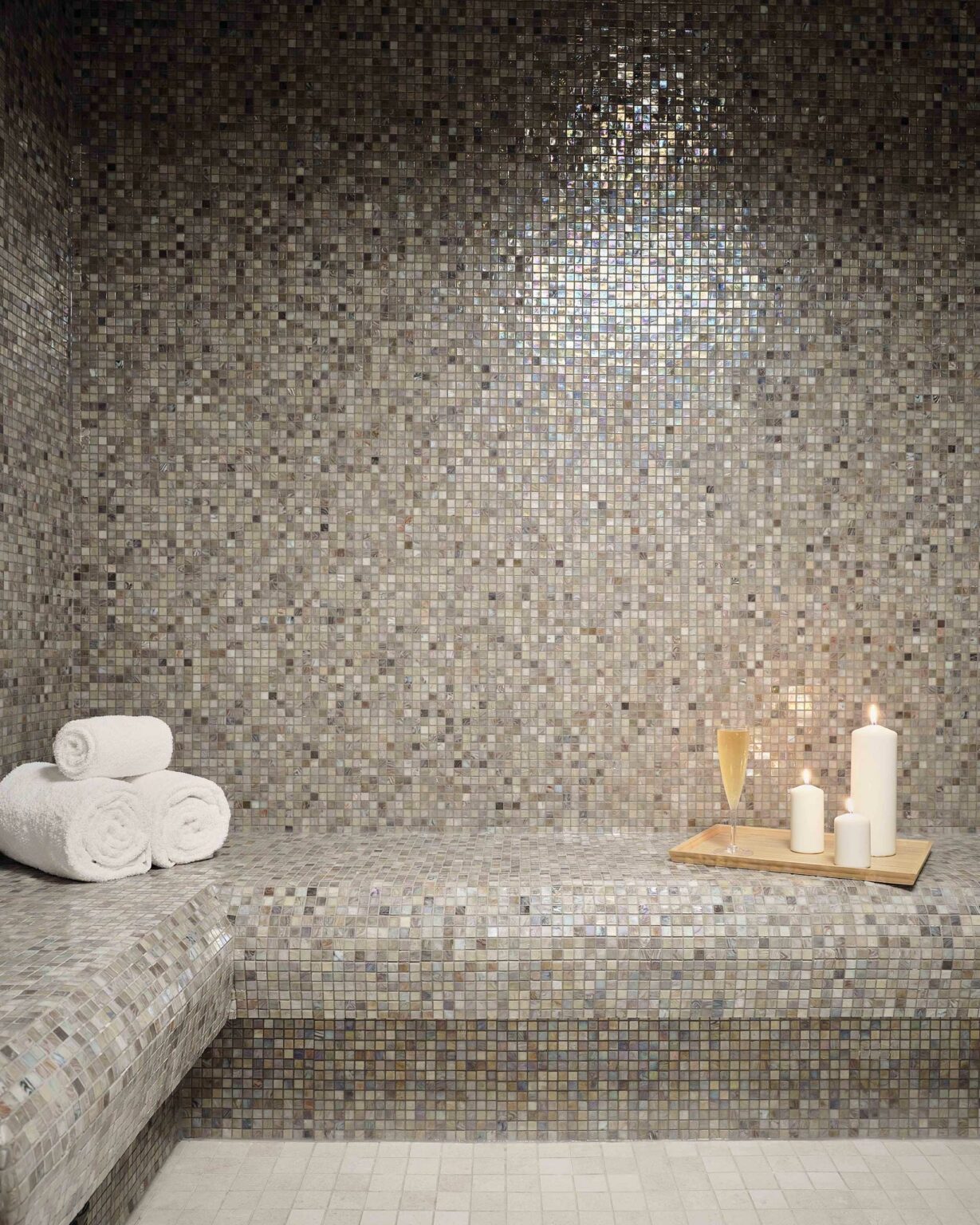 Luxurious spa setting with a mosaic-tiled steam room, rolled white towels, lit candles, and a champagne glass on a wooden tray, creating a serene ambiance.