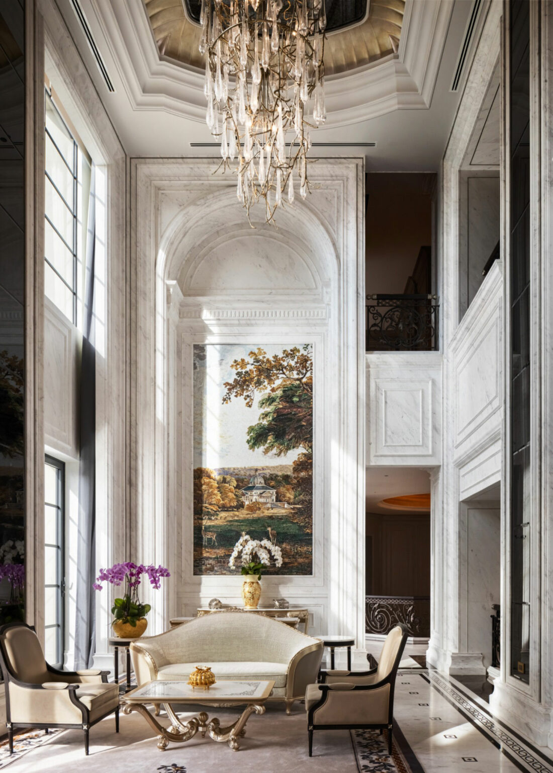 Luxurious living room with tall marble walls, a large landscape painting, elegant chandelier, cream furniture, and purple orchids on a table. Opulent ambiance.