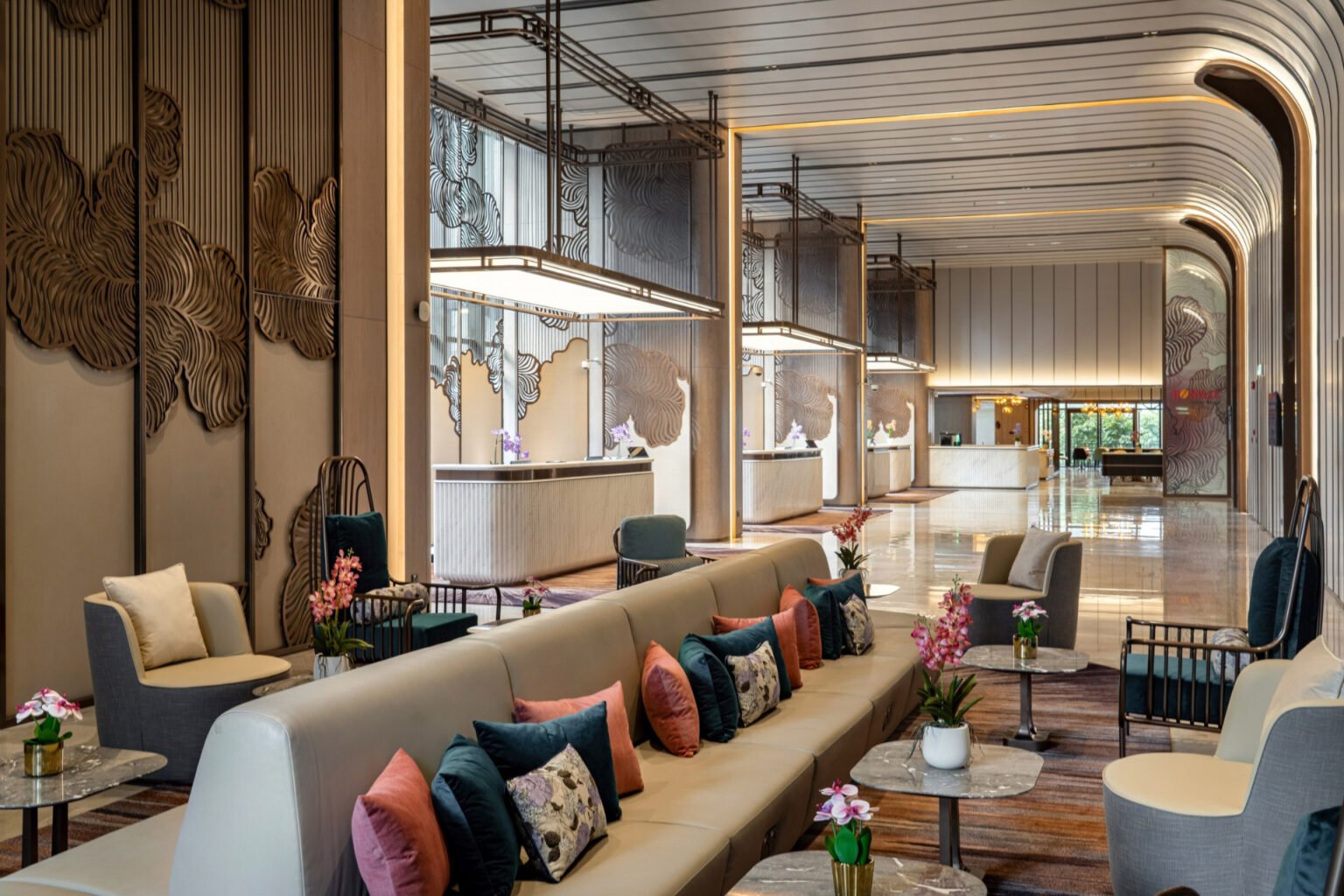 Hotel lobby with high ceilings, modern decor, and floral-themed wall panels. Plush seating with colorful cushions and small tables with flowers create a welcoming atmosphere.