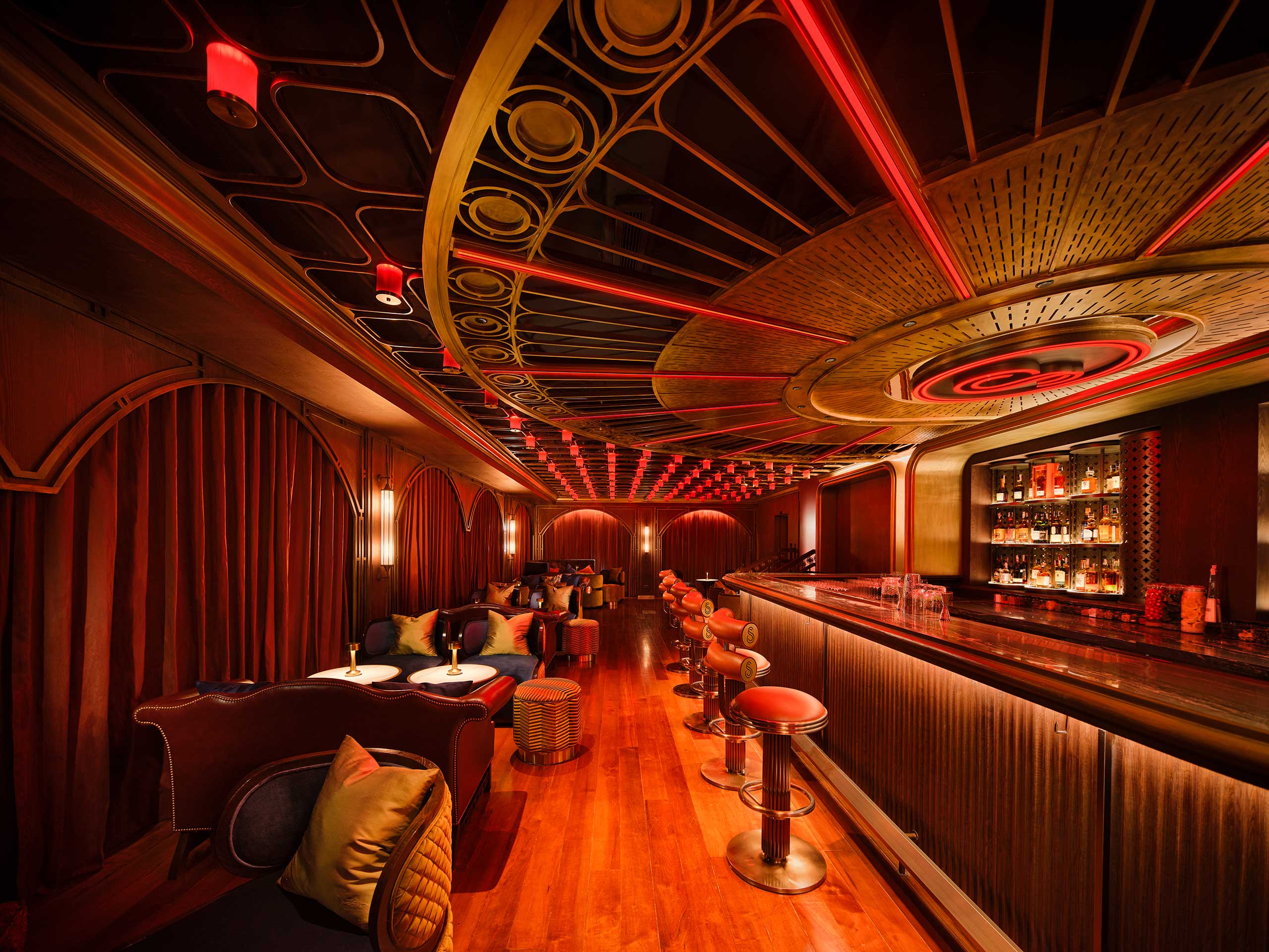 Stylish restaurant bar area with warm lighting, red and gold tones. Curved ceiling with geometric pattern. Cozy seating and a well-stocked bar. Luxurious ambiance.