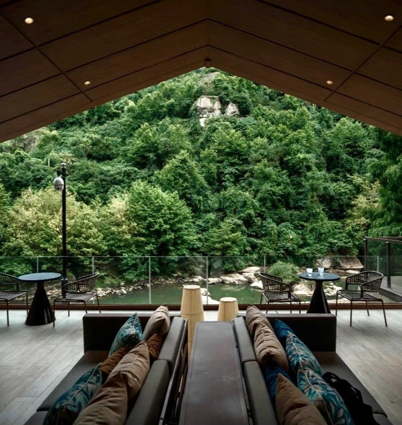 A cozy hotel lounge patio with sofas, patterned cushions, and small tables overlooking a lush green forest and serene river. The ambiance is peaceful and inviting.