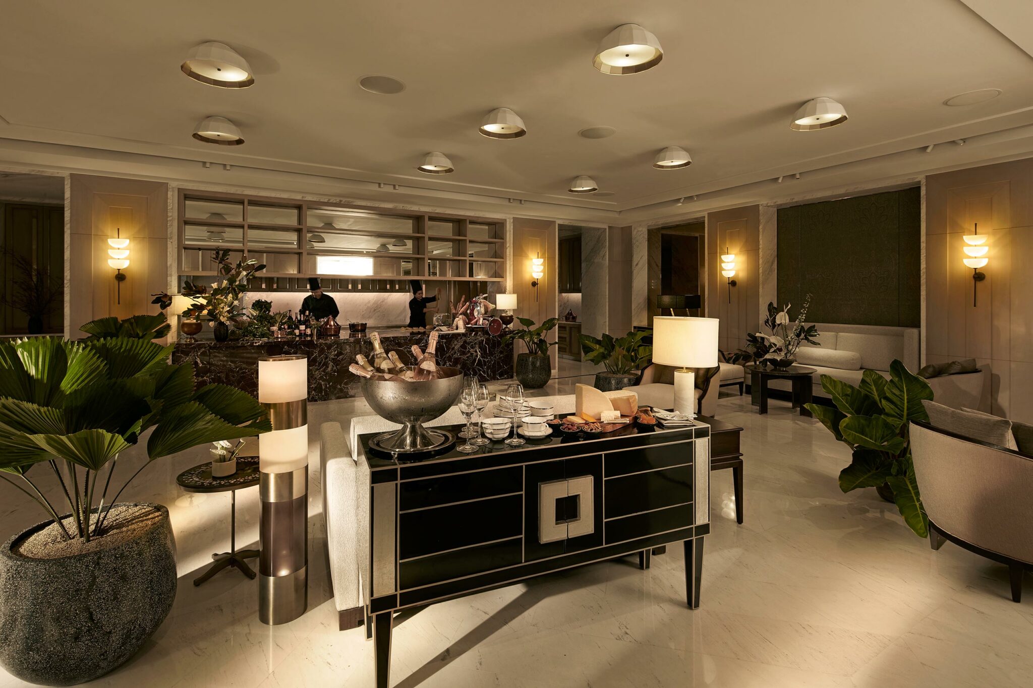 Elegant hotel lounge with warm lighting, featuring a central black sideboard with wine and snacks. Plush seating, large plants, and a stylish bar in the background.